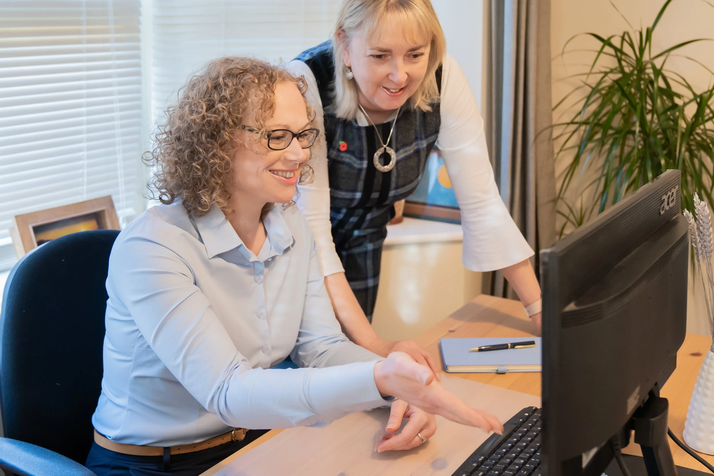 Two women in an office, one sitting at a desk and shaking hands, the other standing and pointing at a computer screen, smiling for the page Expert Bid Writing Service and Bid Support.