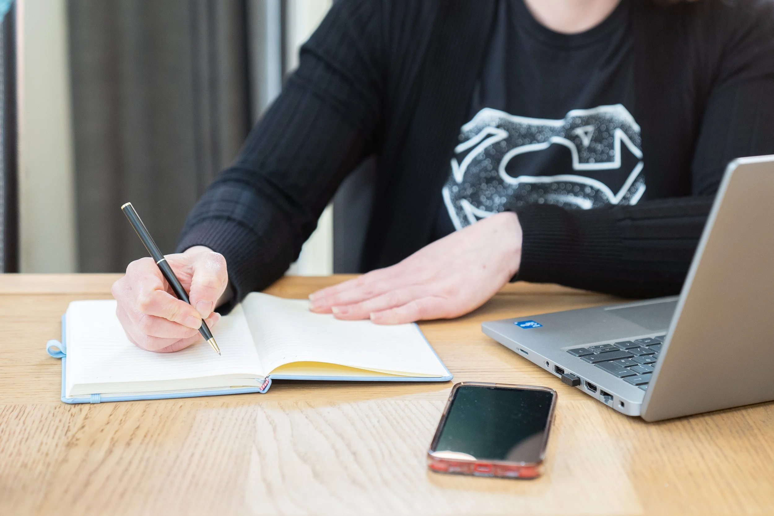 Person writing in a notebook with a pen, sitting at a wooden table with a silver laptop and a smartphone nearby for the page Expert Bid Writing Service and Bid Support.