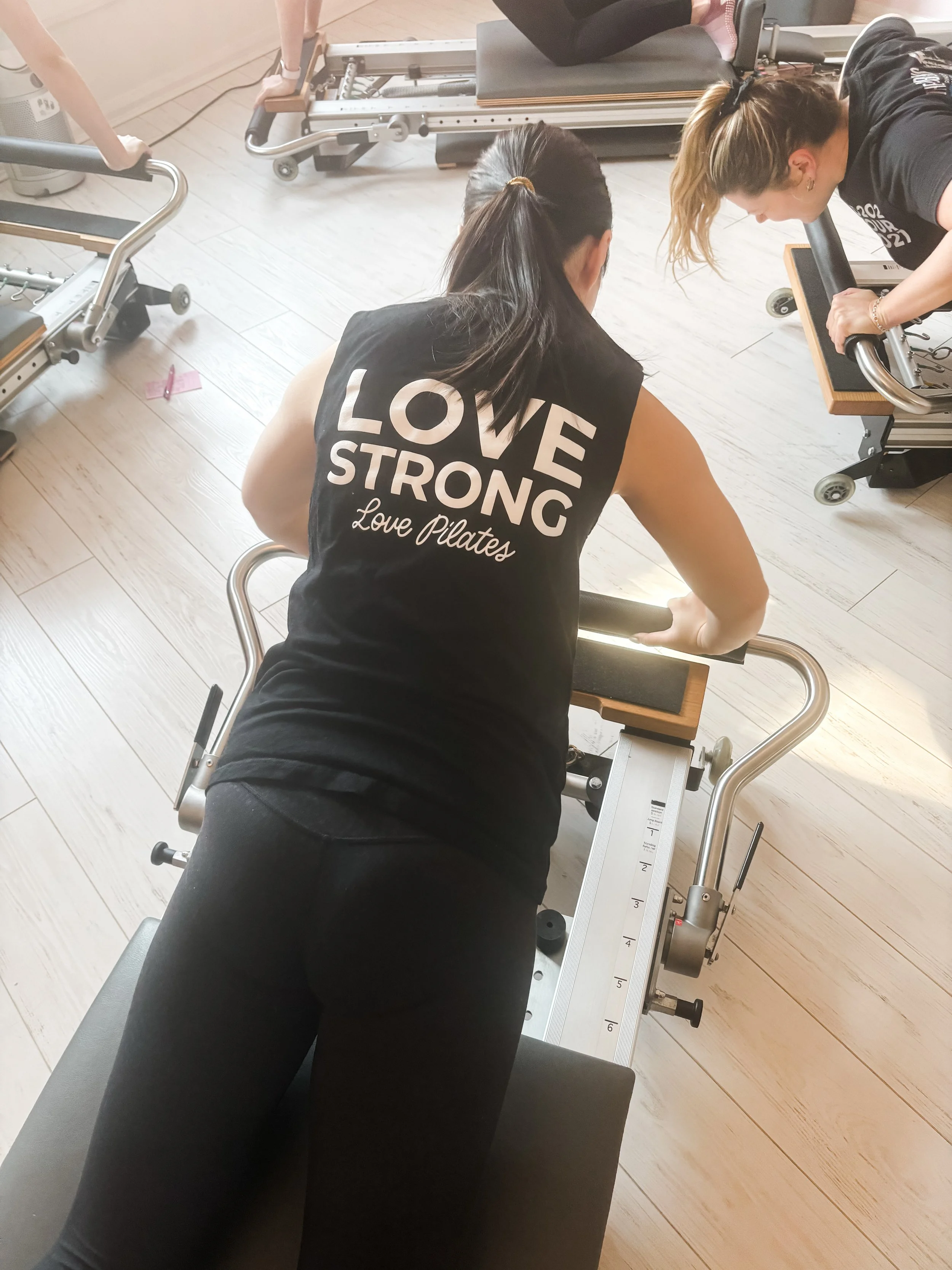 A woman in a black tank top that says 'Love Strong Love Pilates' is kneeling on a Pilates reformer machine, using her arms to push against the foot bar as another person assists her.