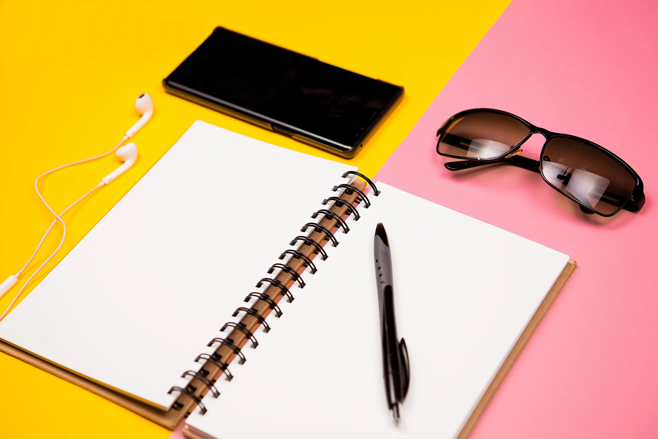 Open spiral notebook with a black pen on top, sunglasses, a black smartphone, and white earphones on a yellow and pink background.