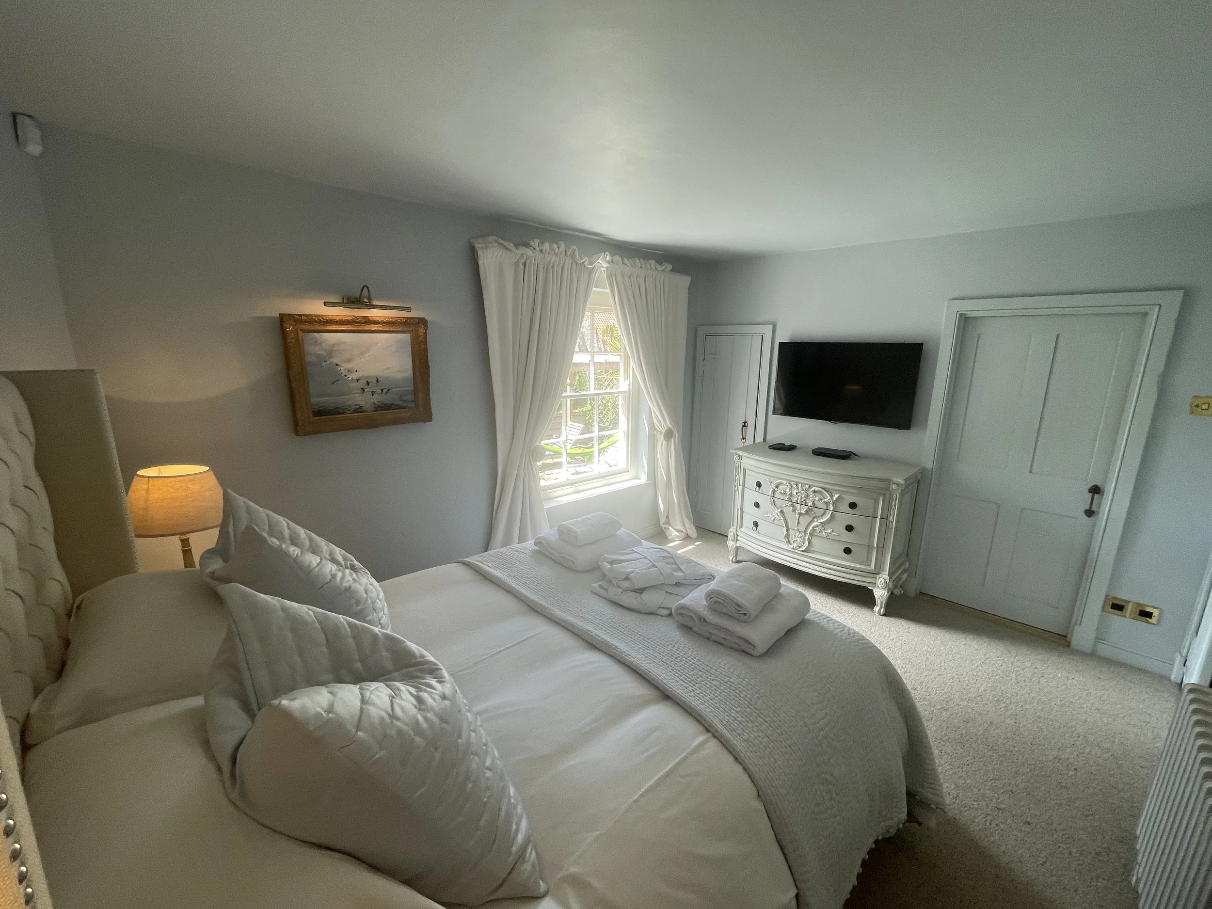 A bedroom with a large bed covered in white quilts and pillows, a nightstand with a lamp, a window with white curtains letting in natural light, a framed wall art, a dresser with decorative details, a flat-screen TV mounted on the wall, and a door in a bedroom with light-colored walls and carpeted floor.