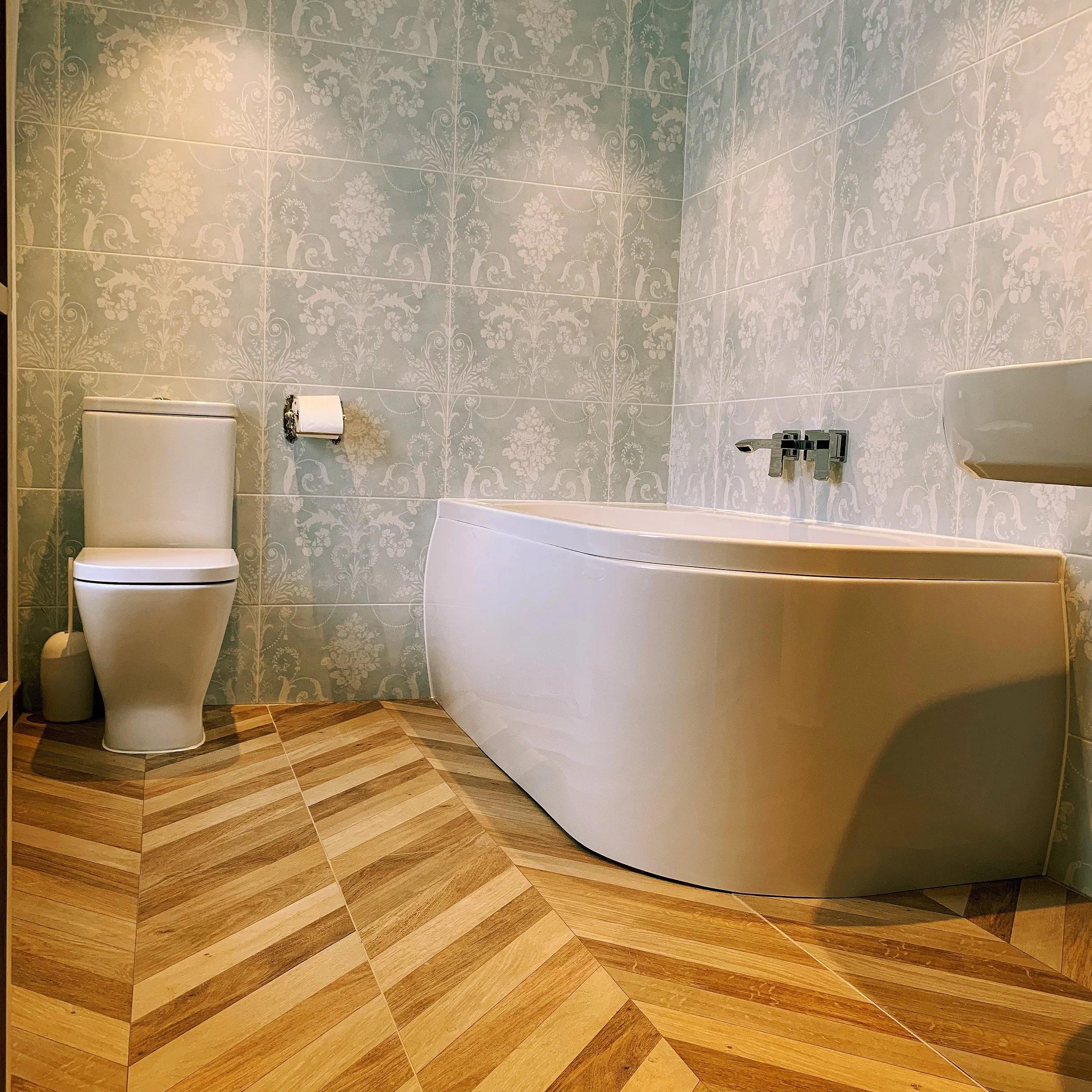 Bathroom with patterned wallpaper, a toilet, and a partial view of a sink and a tub with wood-look tiling on the floor.