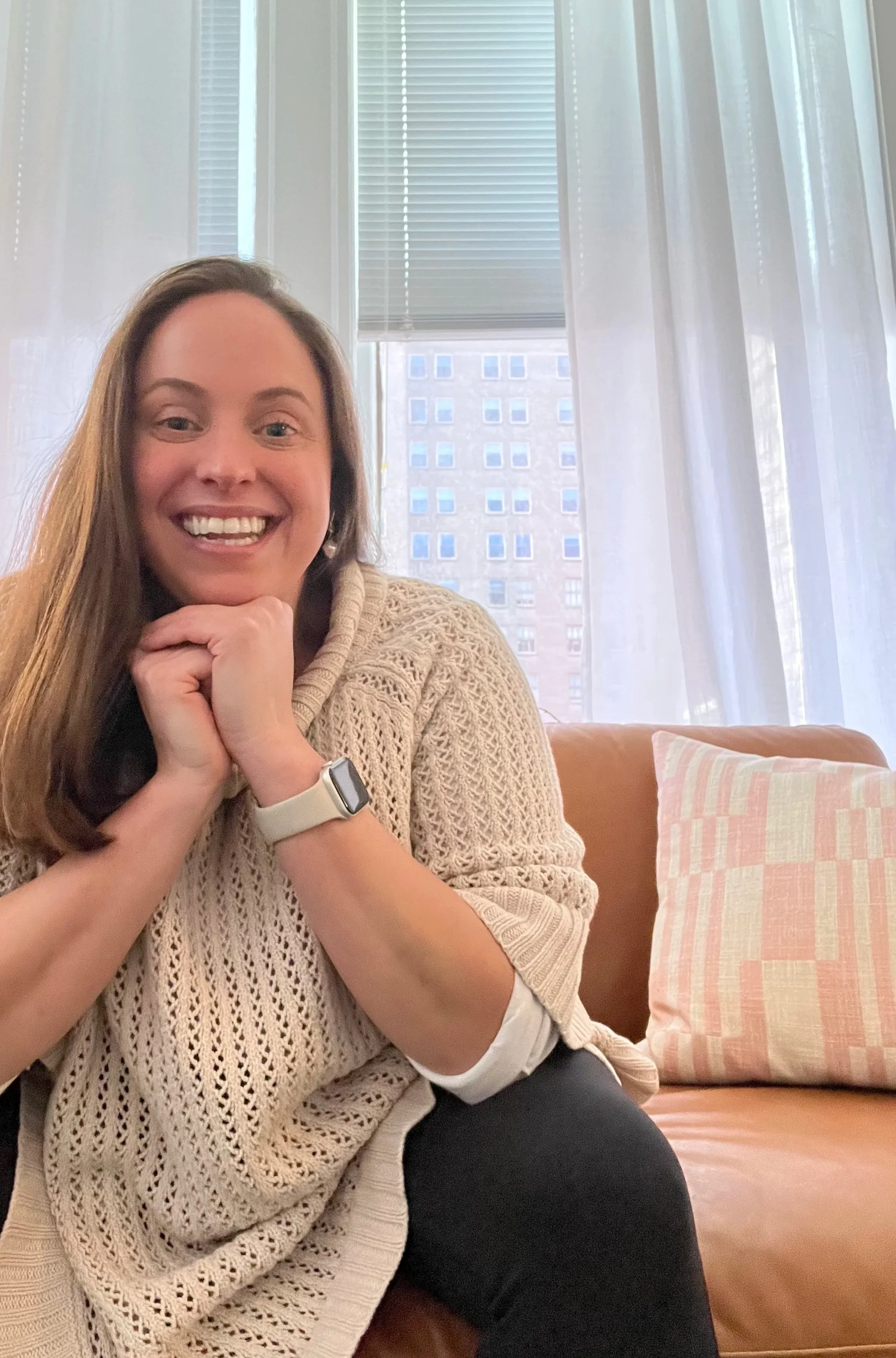 A woman with long brown hair smiling, sitting on a beige couch with her hands under her chin, wearing a cream knitted sweater and an Apple Watch, in a room with large windows and a cityscape view.