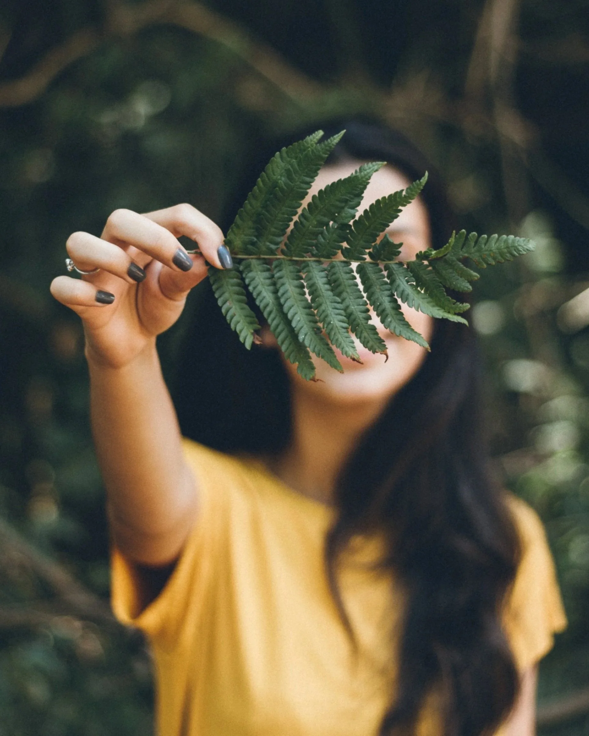 Person in a yellow shirt holding a green fern leaf in front of their face, obscuring their face, in a lush outdoor setting.