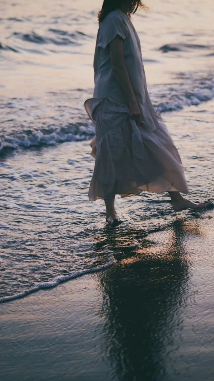 Person standing in shallow ocean water at sunset, wearing a flowing dress.
