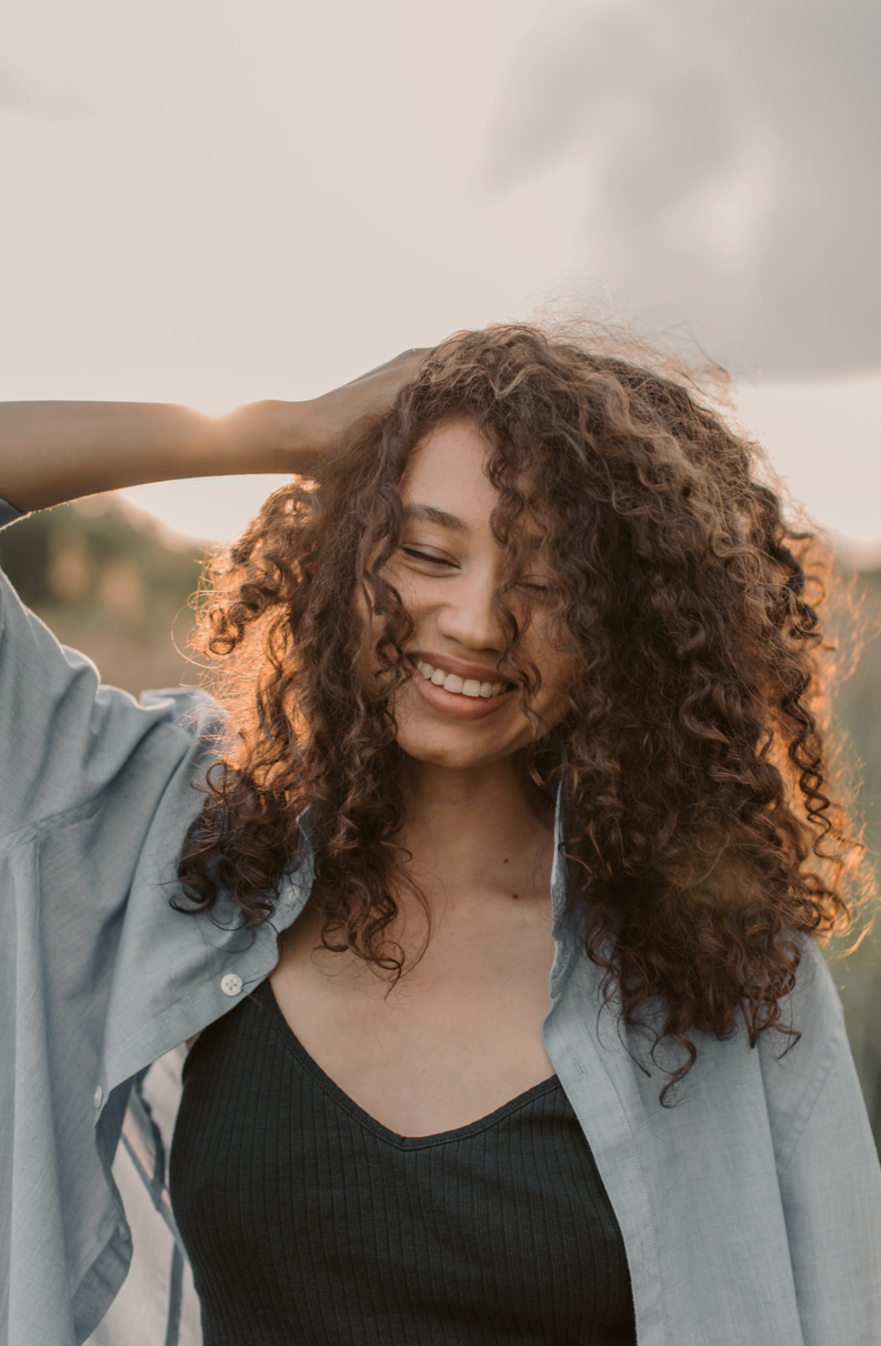 A young woman with curly hair smiling outdoors during sunset, wearing a black top and a light-colored shirt.
