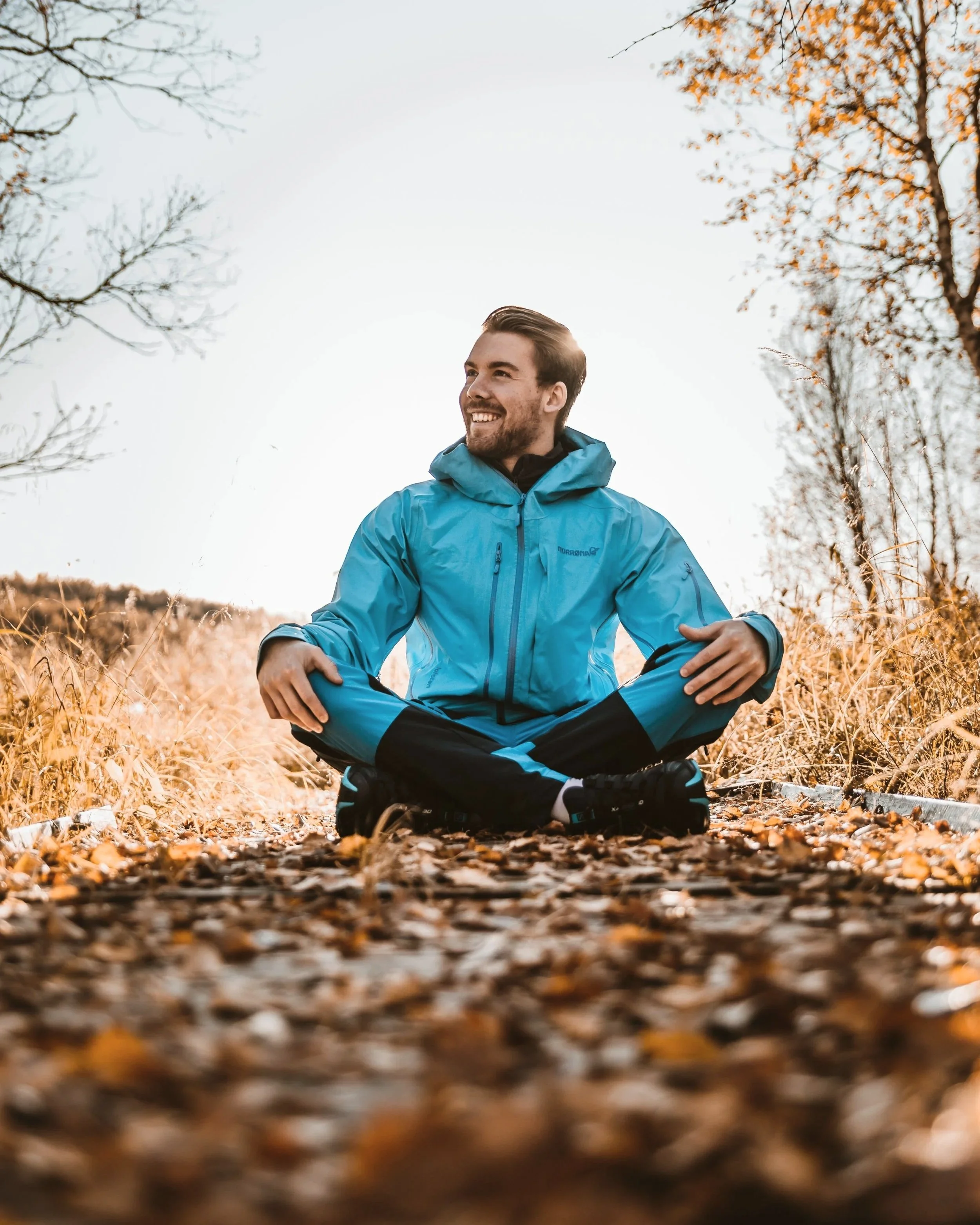 A man sitting cross-legged outdoors on a leaf-covered path in a park during autumn, smiling and wearing a blue waterproof jacket.