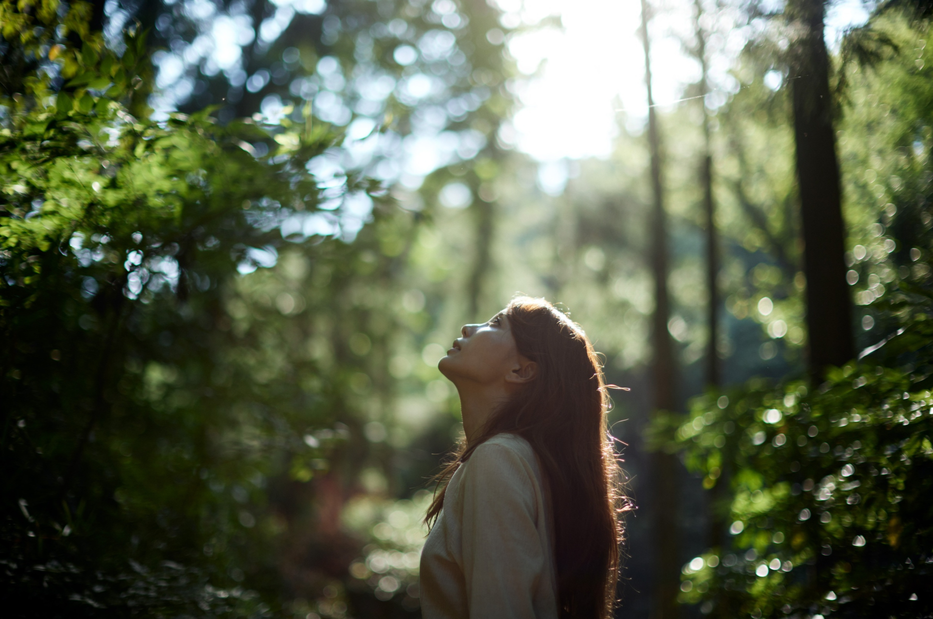 A woman in a beige jacket standing in a forest with sunlight shining through trees, looking up with her head tilted back.