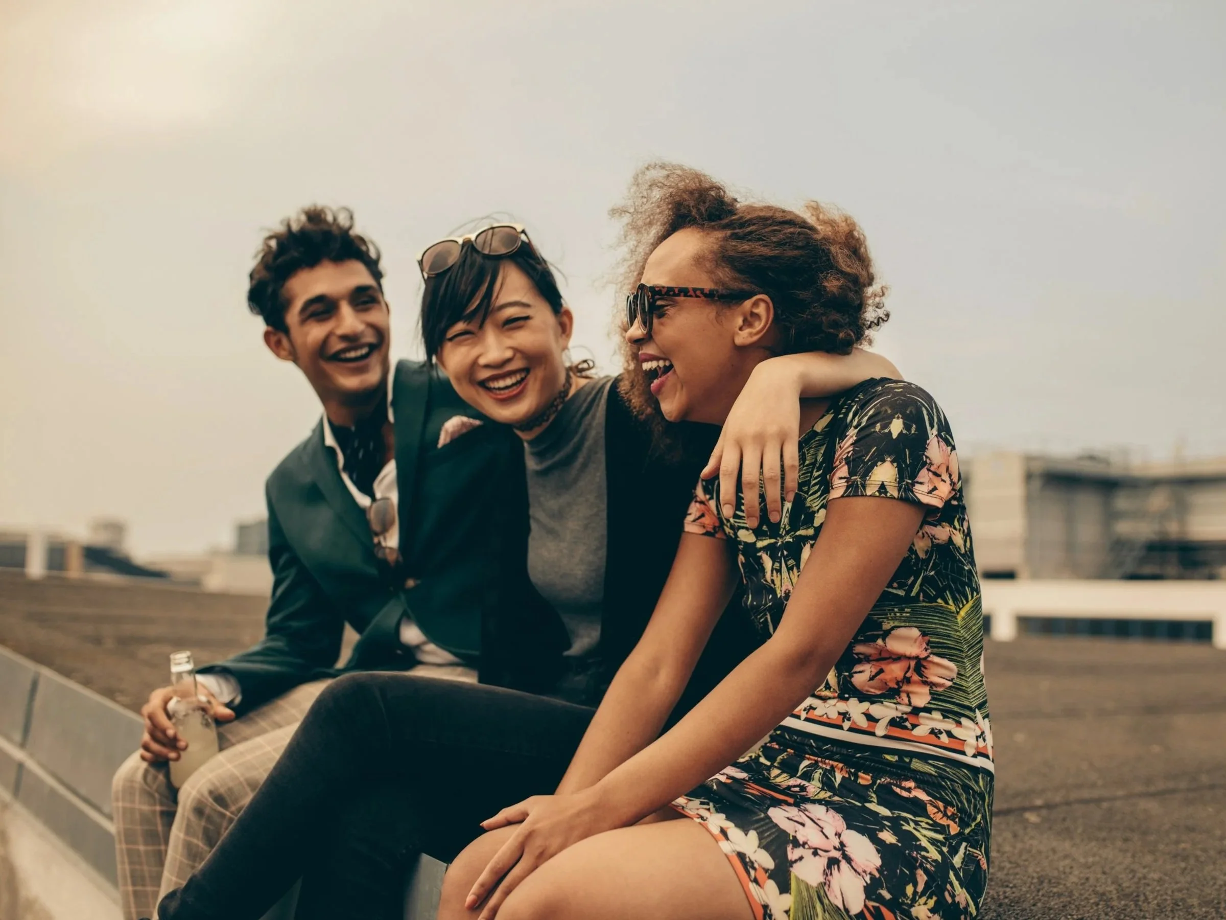 Three friends sitting on a rooftop, smiling and laughing together, enjoying each other's company on a cloudy day.