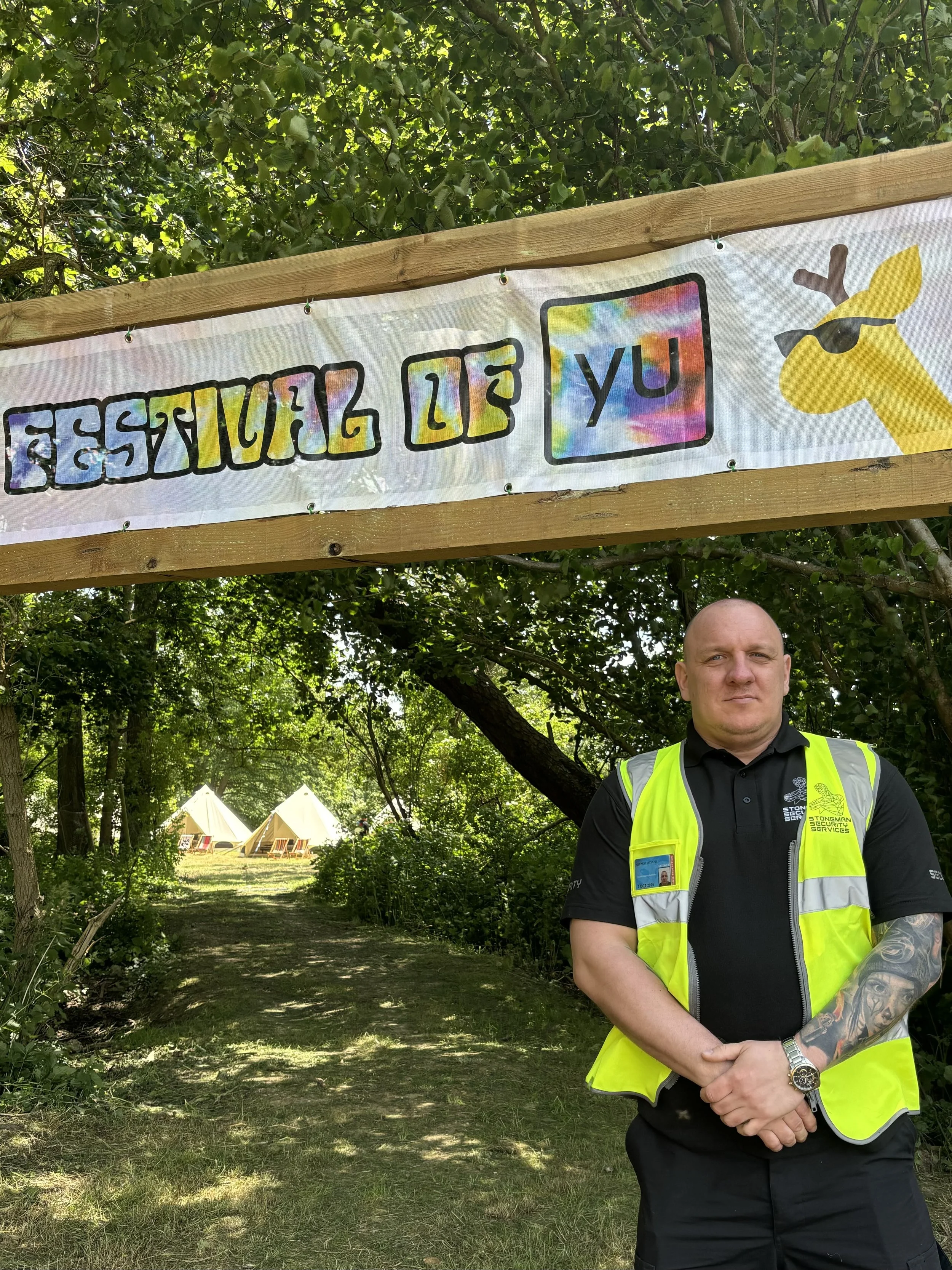 Security guard standing beside a sign reading 'Festival of Yu' at an outdoor event with tents in the background.
