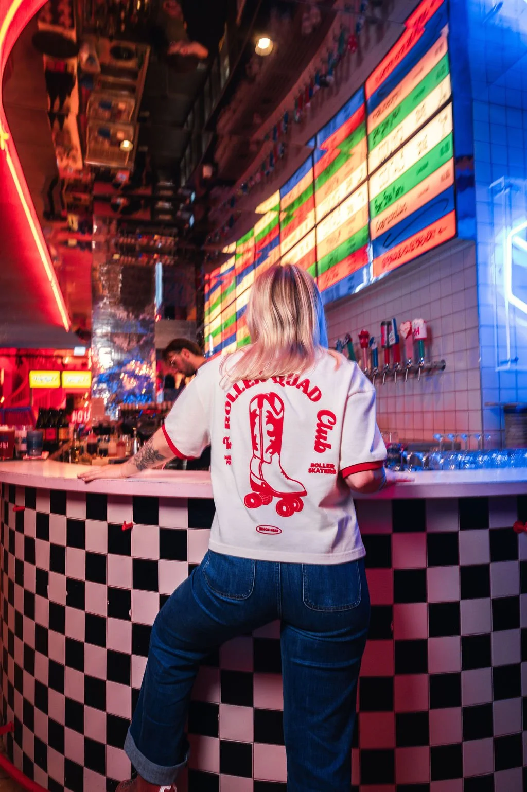 Une jeune femme se tient sur un trottoir en ville, portant un sweat gris avec l'inscription "ROLLER" en jaune, une casquette grise, et un collier de coquillages, avec des palmiers et des bâtiments modernes en arrière-plan.
