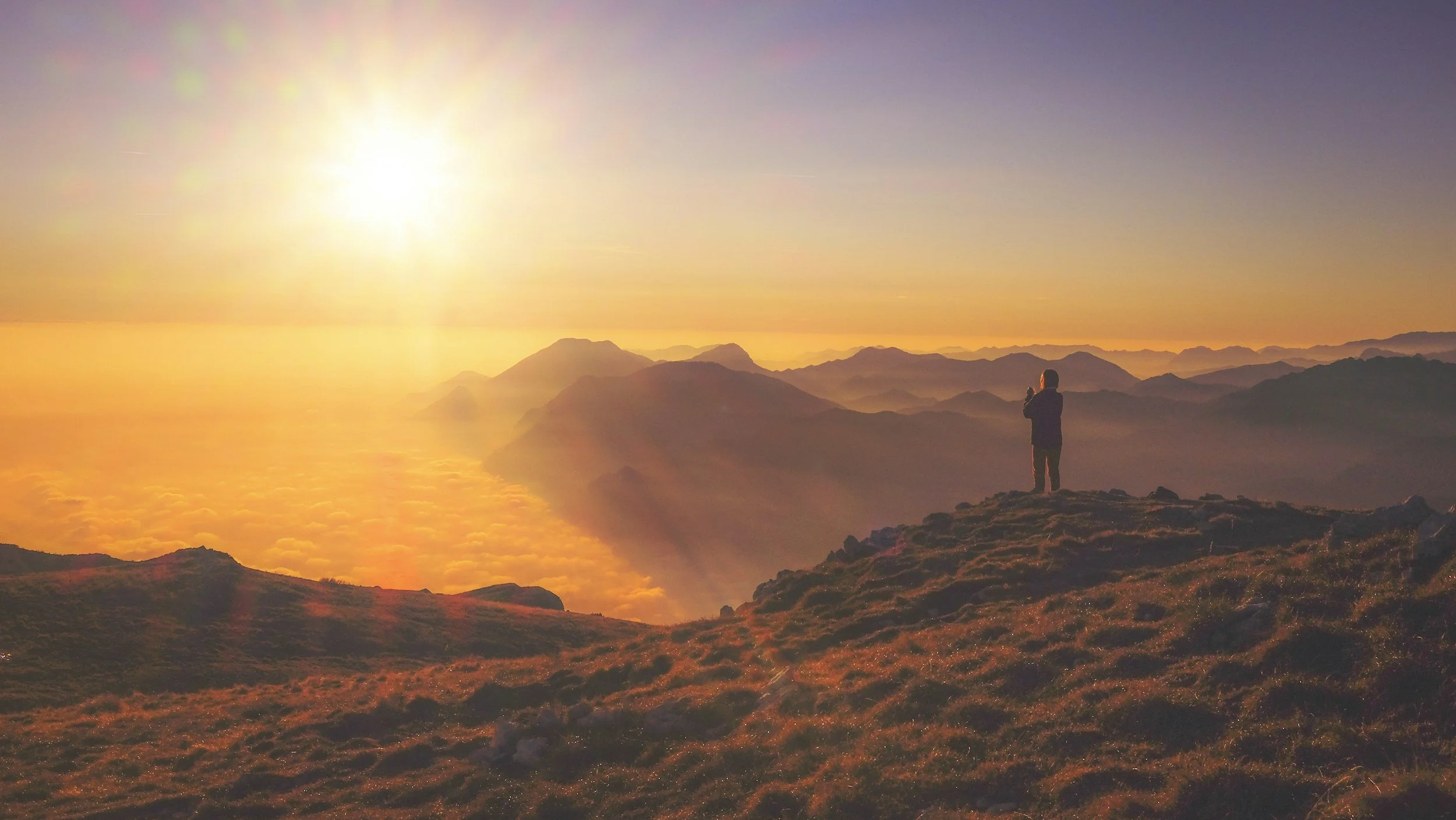 Silhouette of a person standing on a grassy hilltop during a vibrant sunrise, with mist-covered mountains and a golden sky in the background.