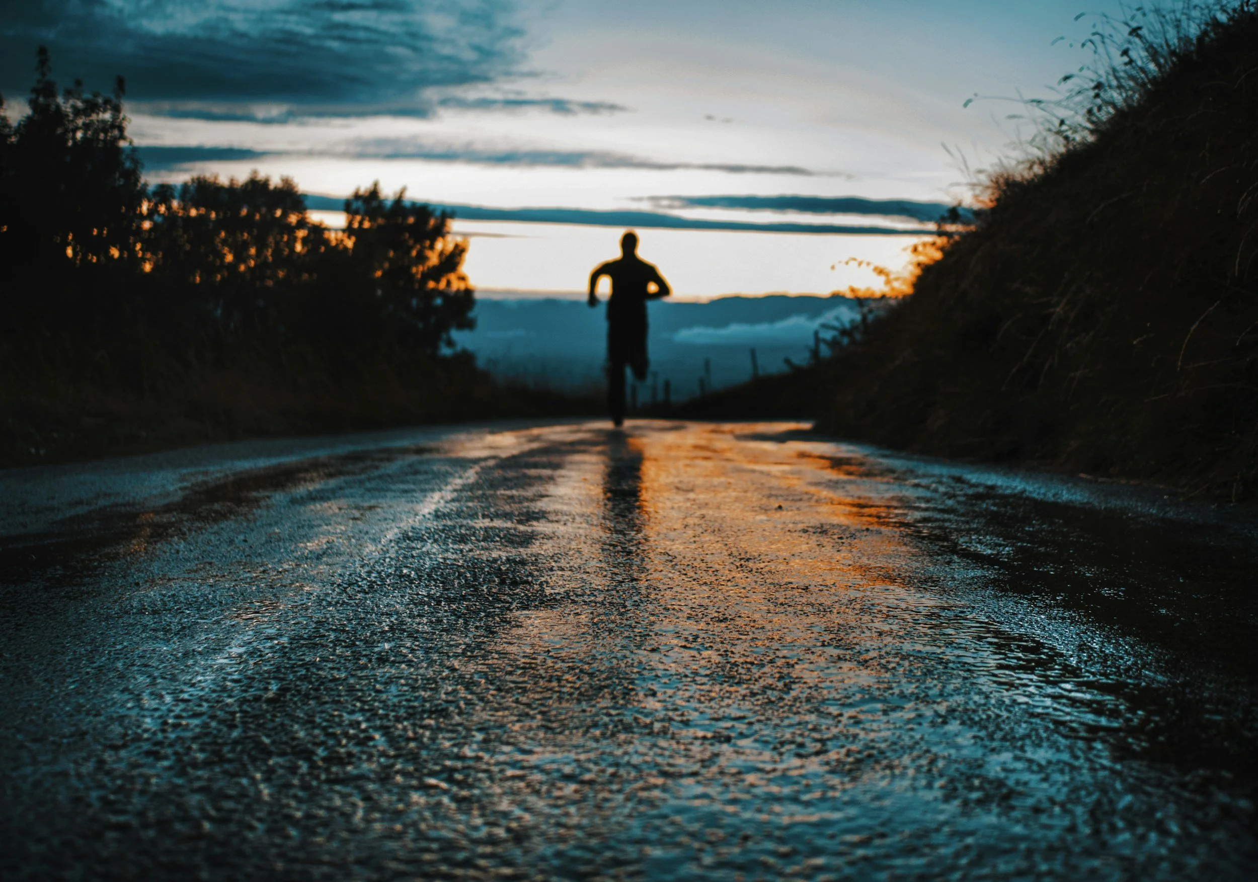 Silhouette of a person running on a wet road at sunset, with trees on the side and a cloudy sky.