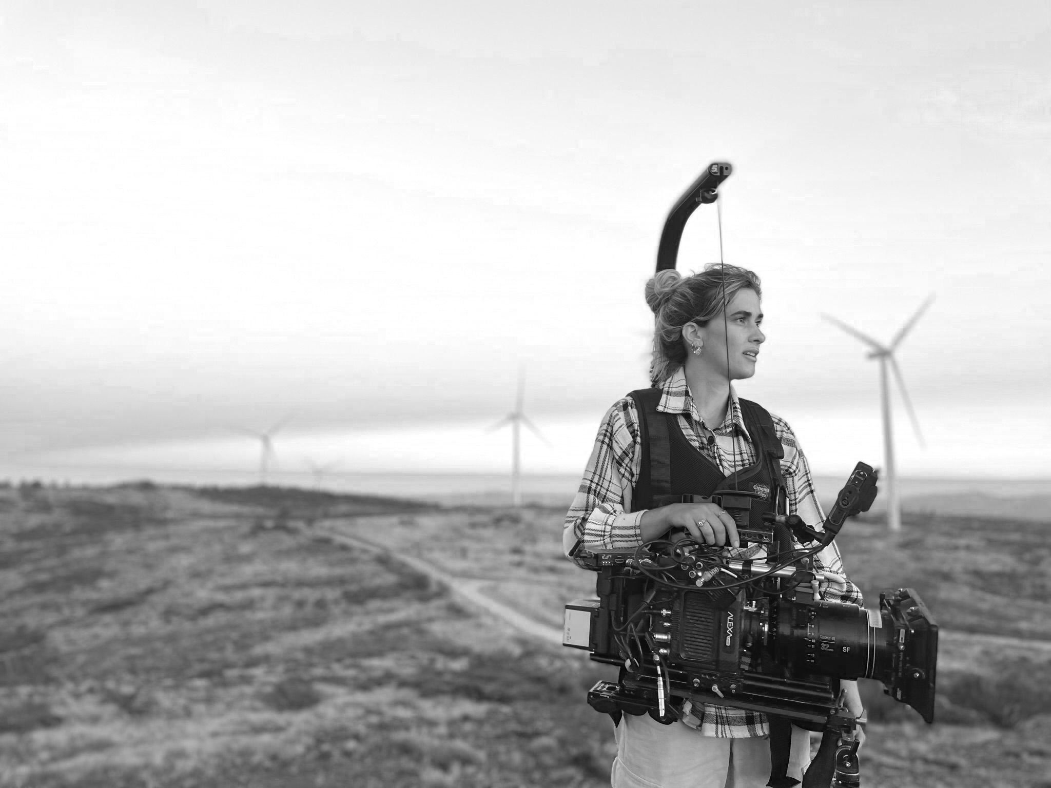 Black and white photo of a woman standing outdoors in a rural landscape with wind turbines in the background, holding a professional video camera.