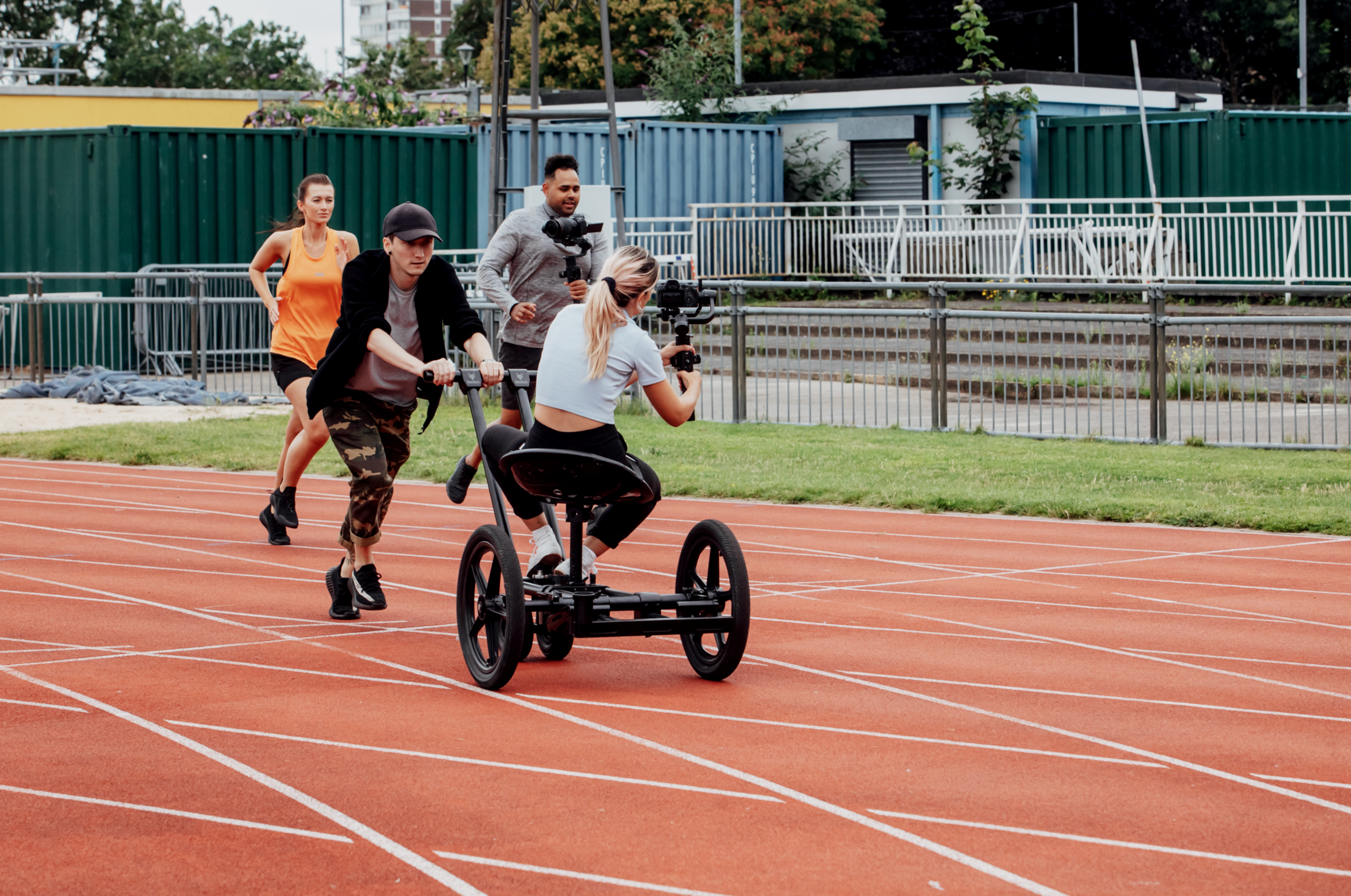 Woman in a racing wheelchair being pushed by a person on a running track, with two cameramen filming the scene, and another woman jogging in the background.