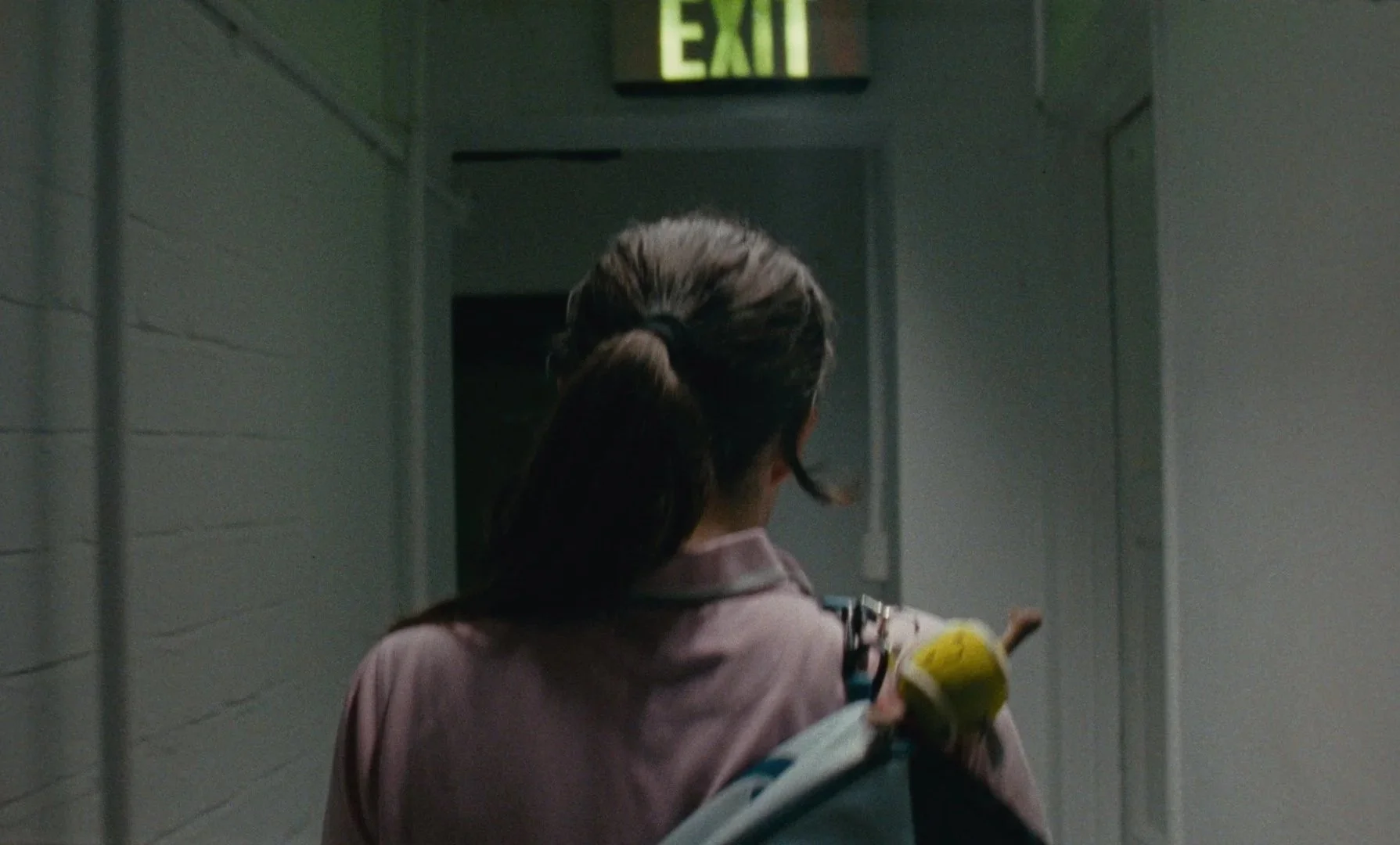 Person with a ponytail holding a yellow stuffed toy, walking down a corridor with an illuminated exit sign above.