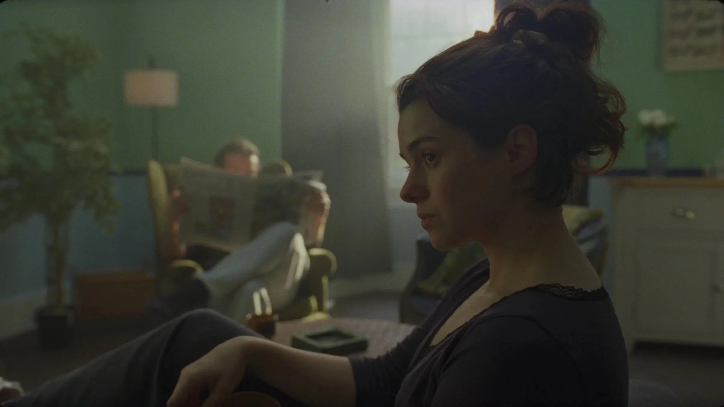 A woman with short curly hair sitting in a dimly lit room, looking to her left, with a man reading a newspaper in the background.