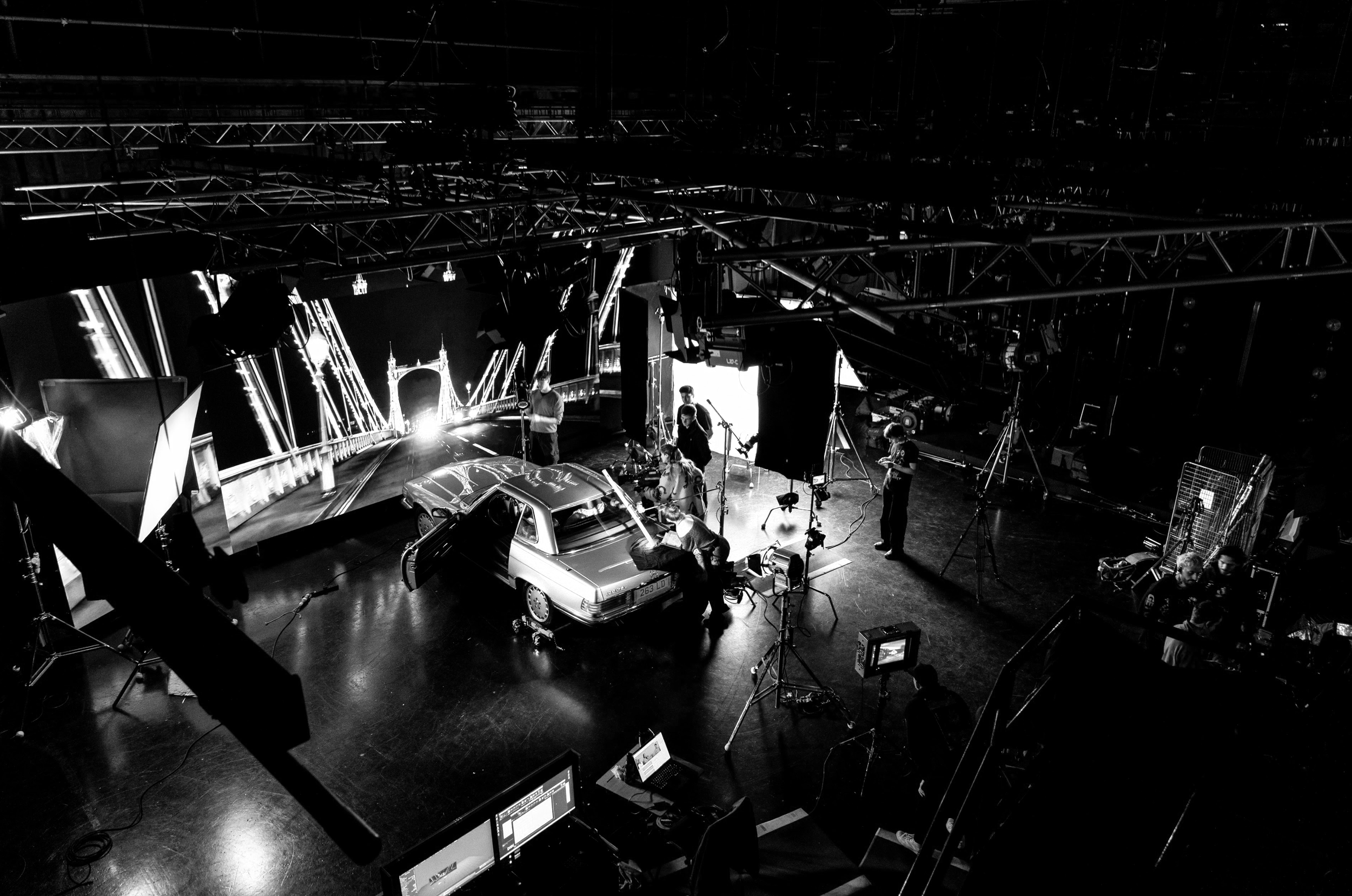 A behind-the-scenes view of a film or photography studio with a vintage car on a set, crew members working, and filming equipment including cameras, lights, and monitors.