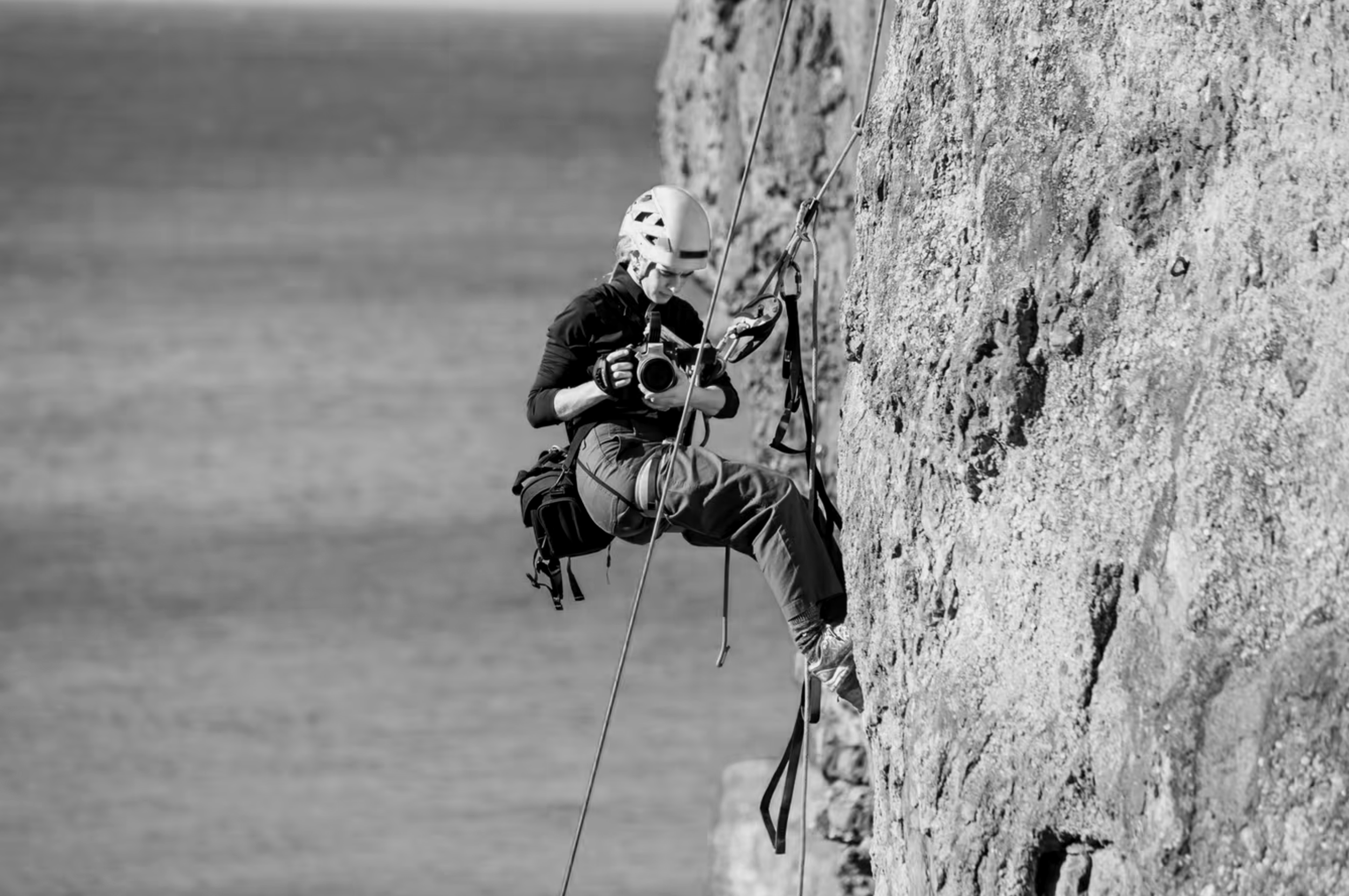 A person climbing a rock face near water, wearing a pink helmet, black jacket, and gray pants, holding a camera in one hand, with climbing gear attached to their harness.