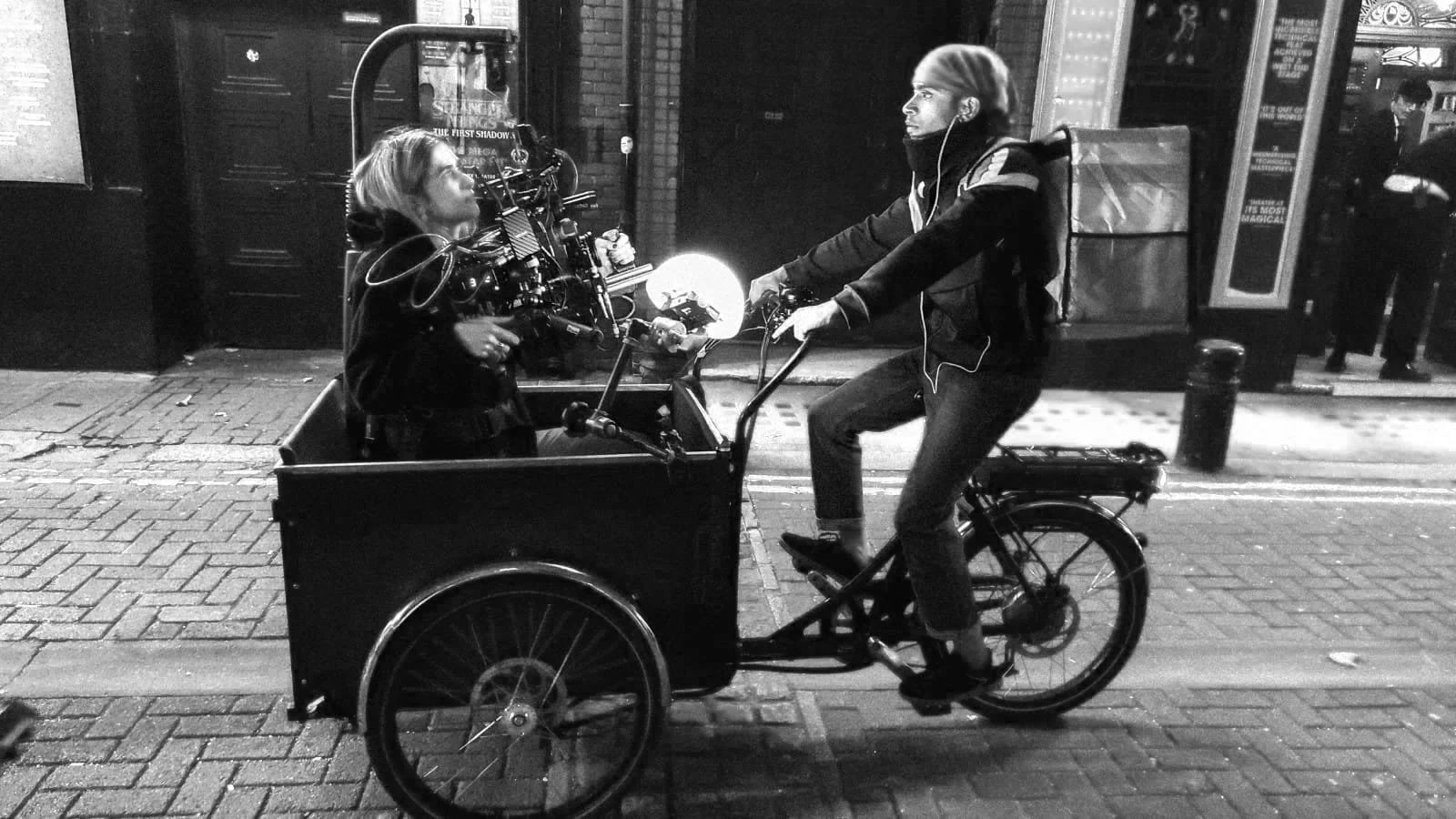 A man riding a bicycle with a cargo hold in the front is talking to a woman sitting inside the cargo area. The scene is lit with red and orange streetlights.