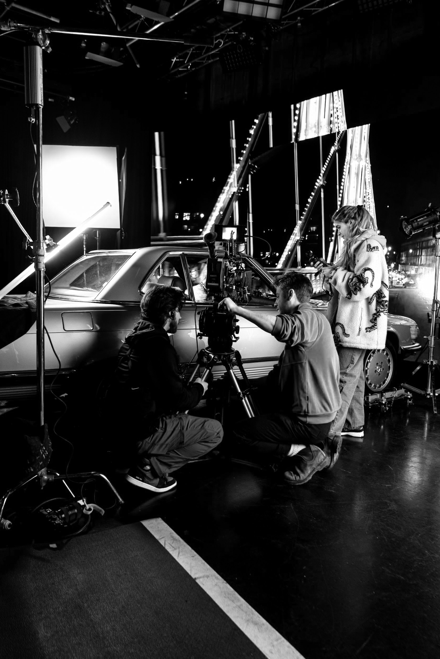 Filming scene with a vintage car on a night-lit set, crew members adjusting equipment and a woman looking at her phone.