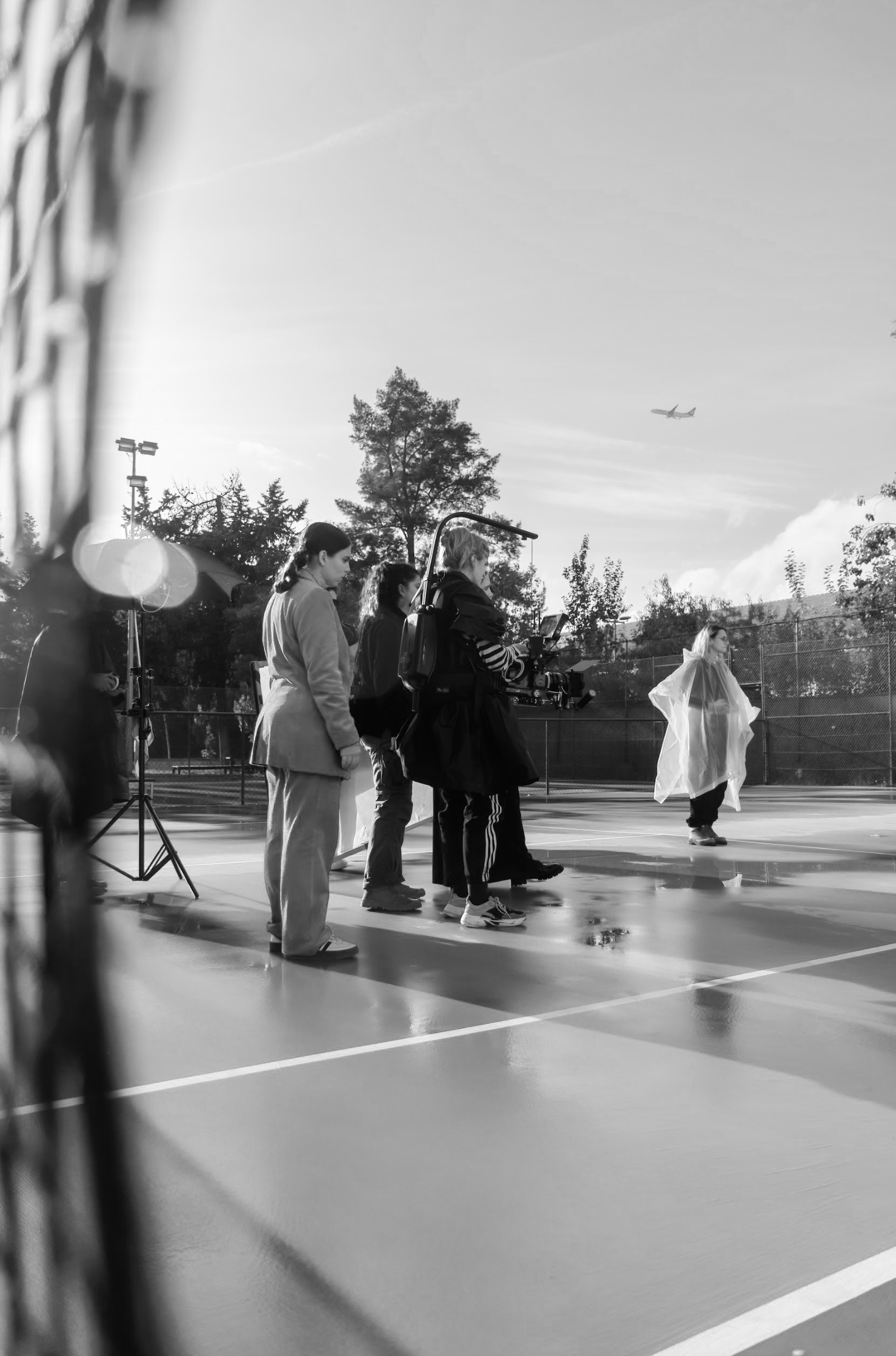 Film crew filming on an outdoor tennis court with a woman in a white raincoat, trees, and airplane overhead.
