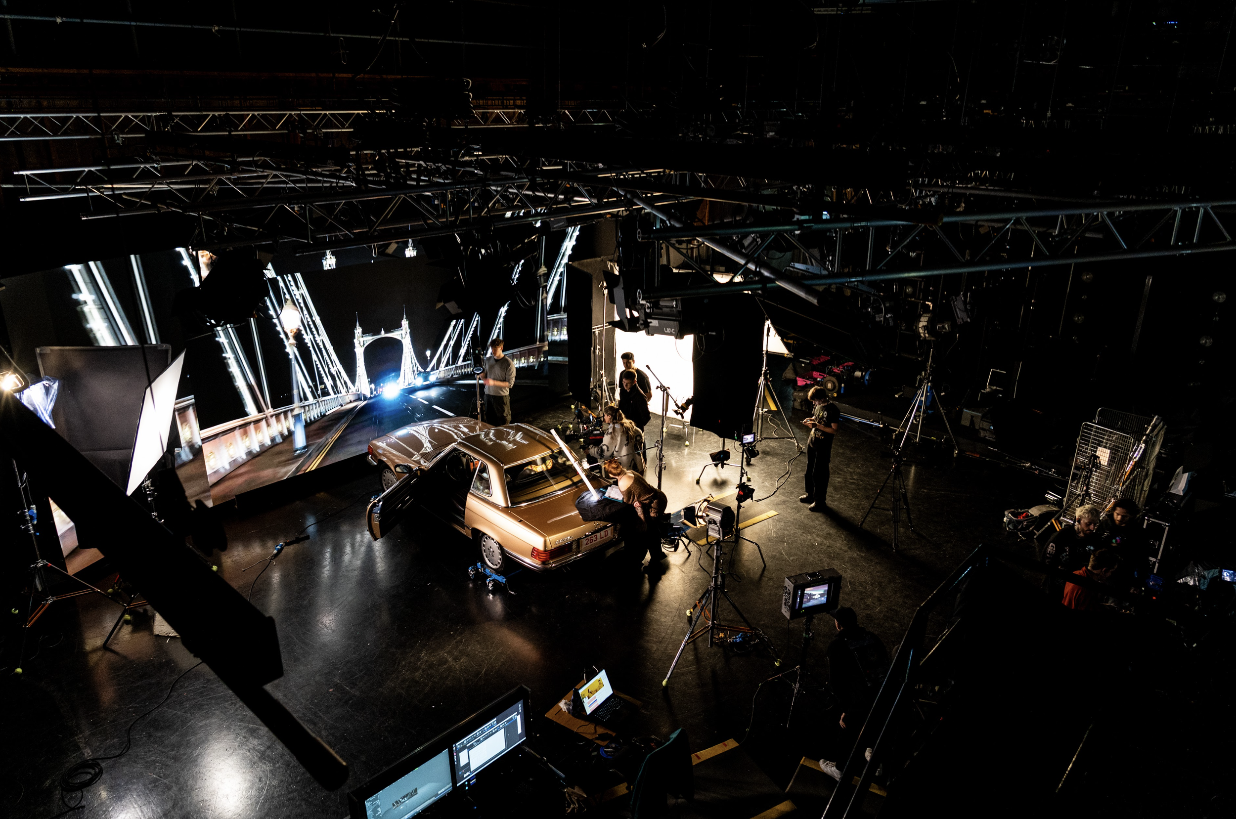 A behind-the-scenes view of a film or photography studio with a vintage car on a set, crew members working, and filming equipment including cameras, lights, and monitors.