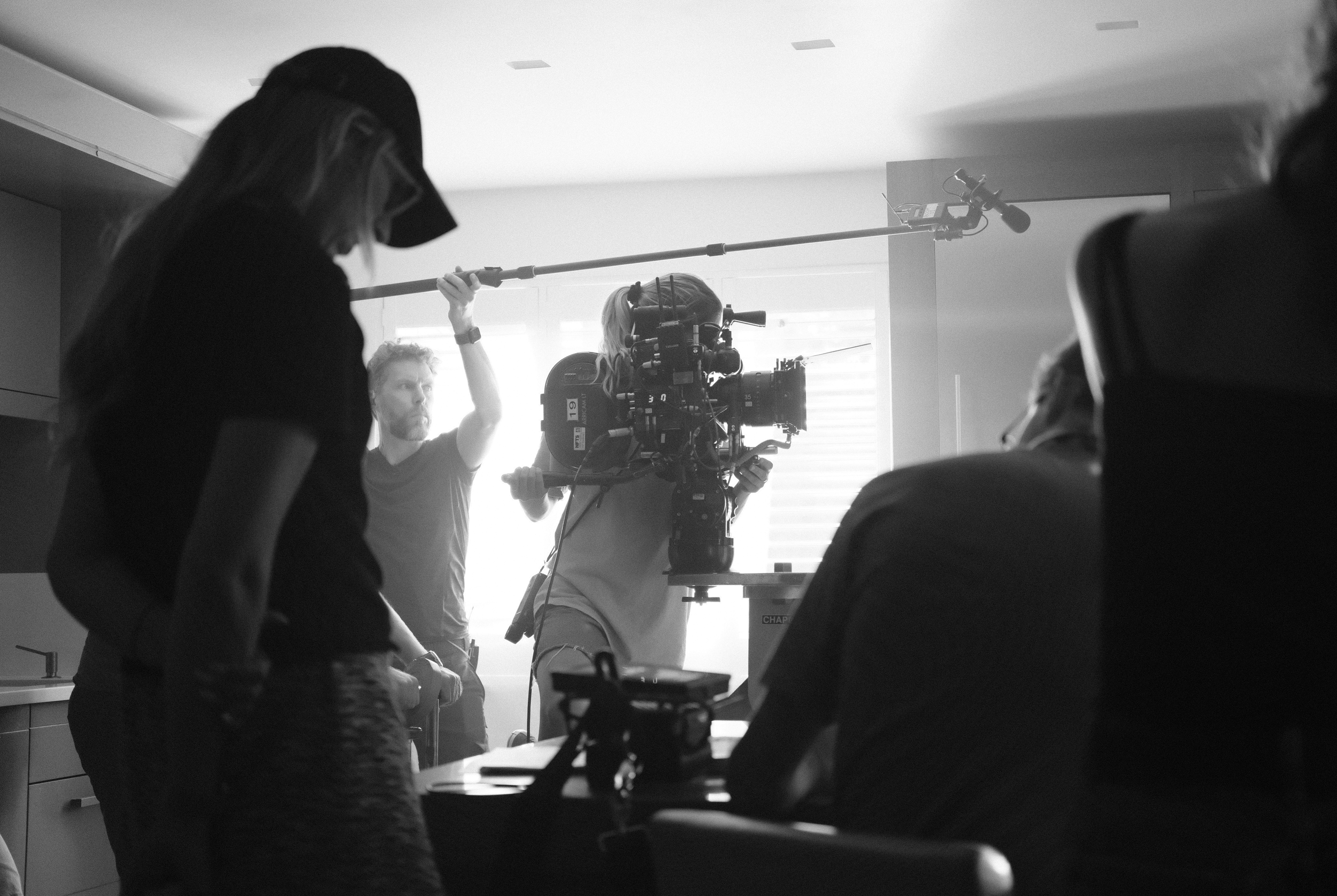 Filmmaking crew working on a set with camera and lighting equipment in a room with windows.