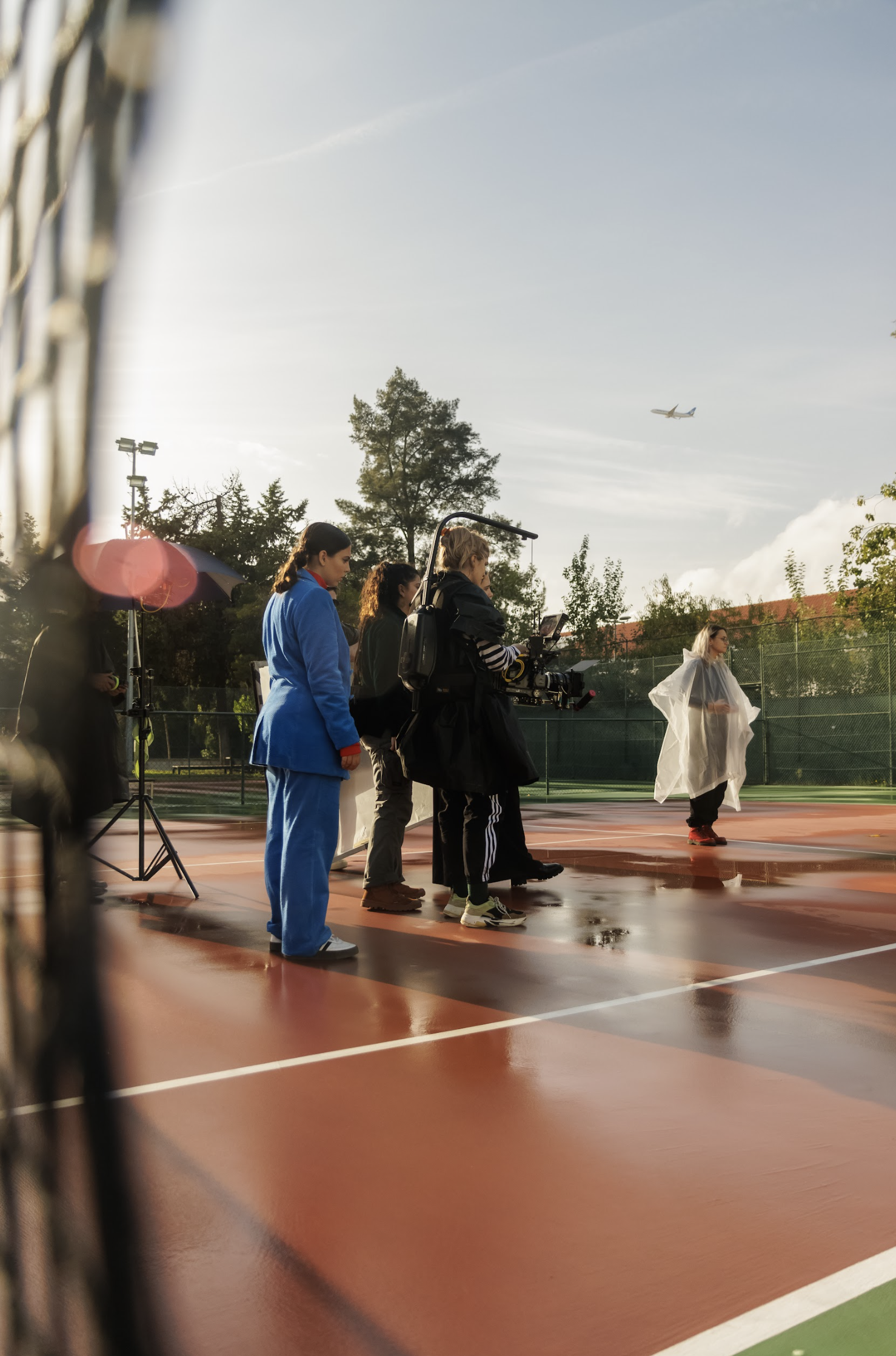 Film crew filming on an outdoor tennis court with a woman in a white raincoat, trees, and airplane overhead.