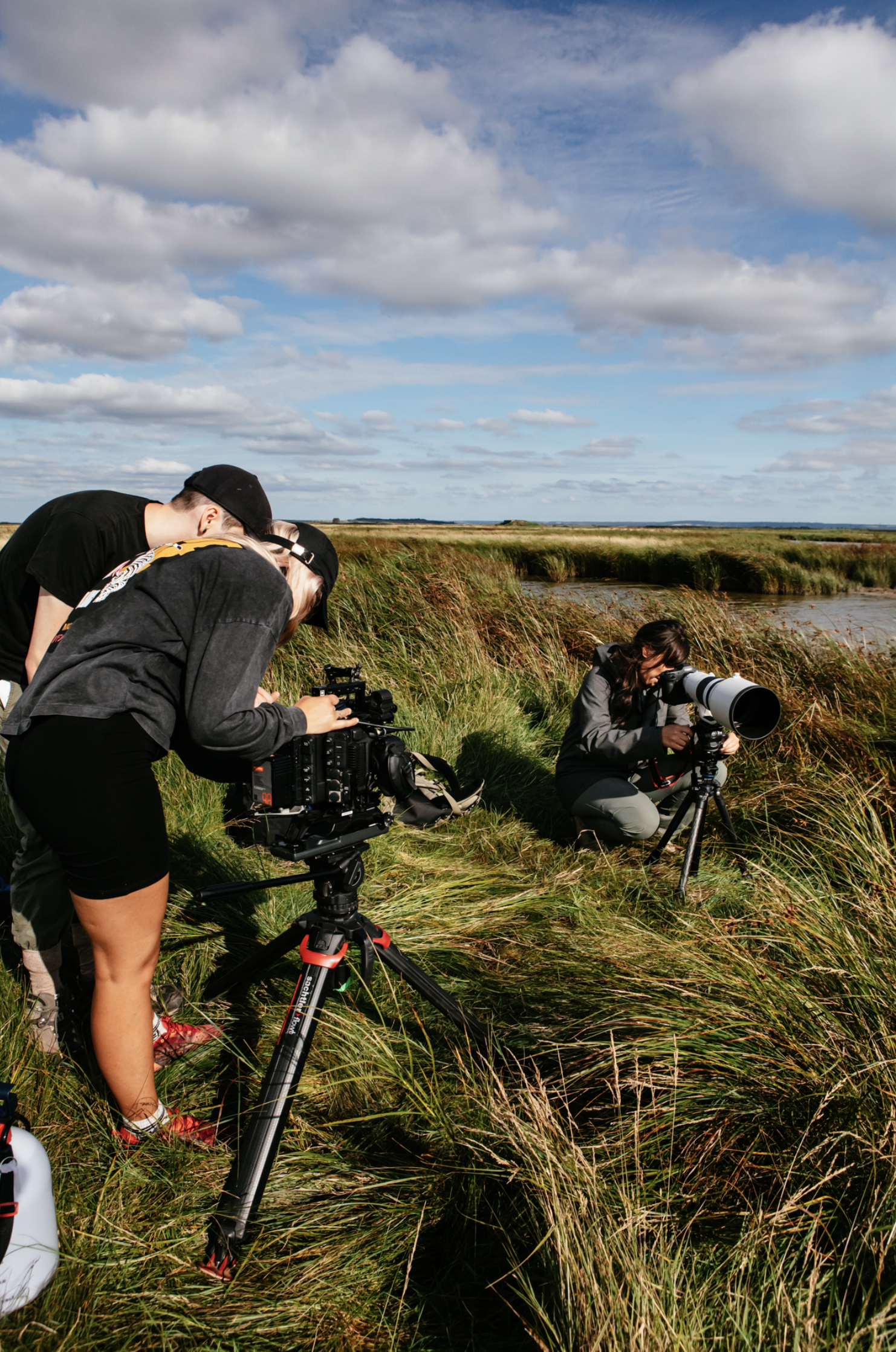 Group of three people filming nature scene with cameras on tripods in a grassy marshland under a partly cloudy sky.