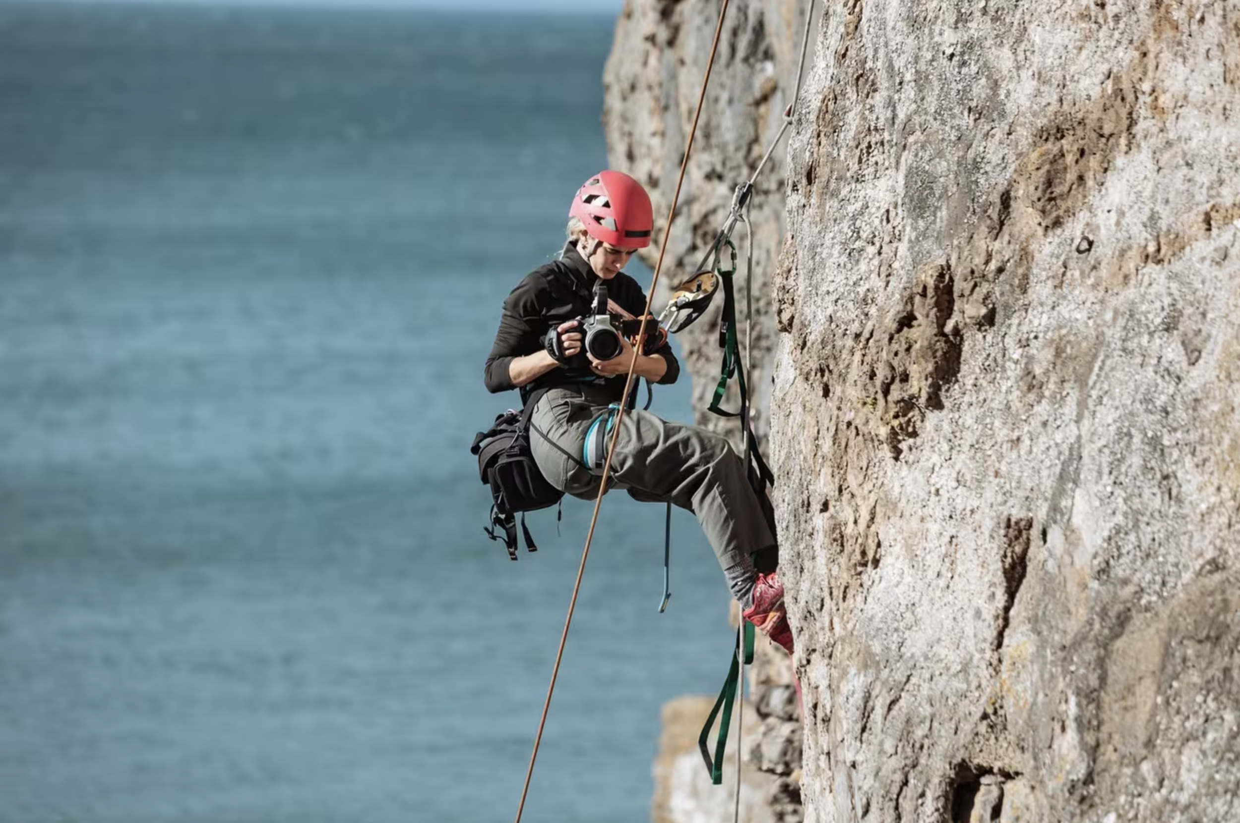 A person climbing a rock face near water, wearing a pink helmet, black jacket, and gray pants, holding a camera in one hand, with climbing gear attached to their harness.