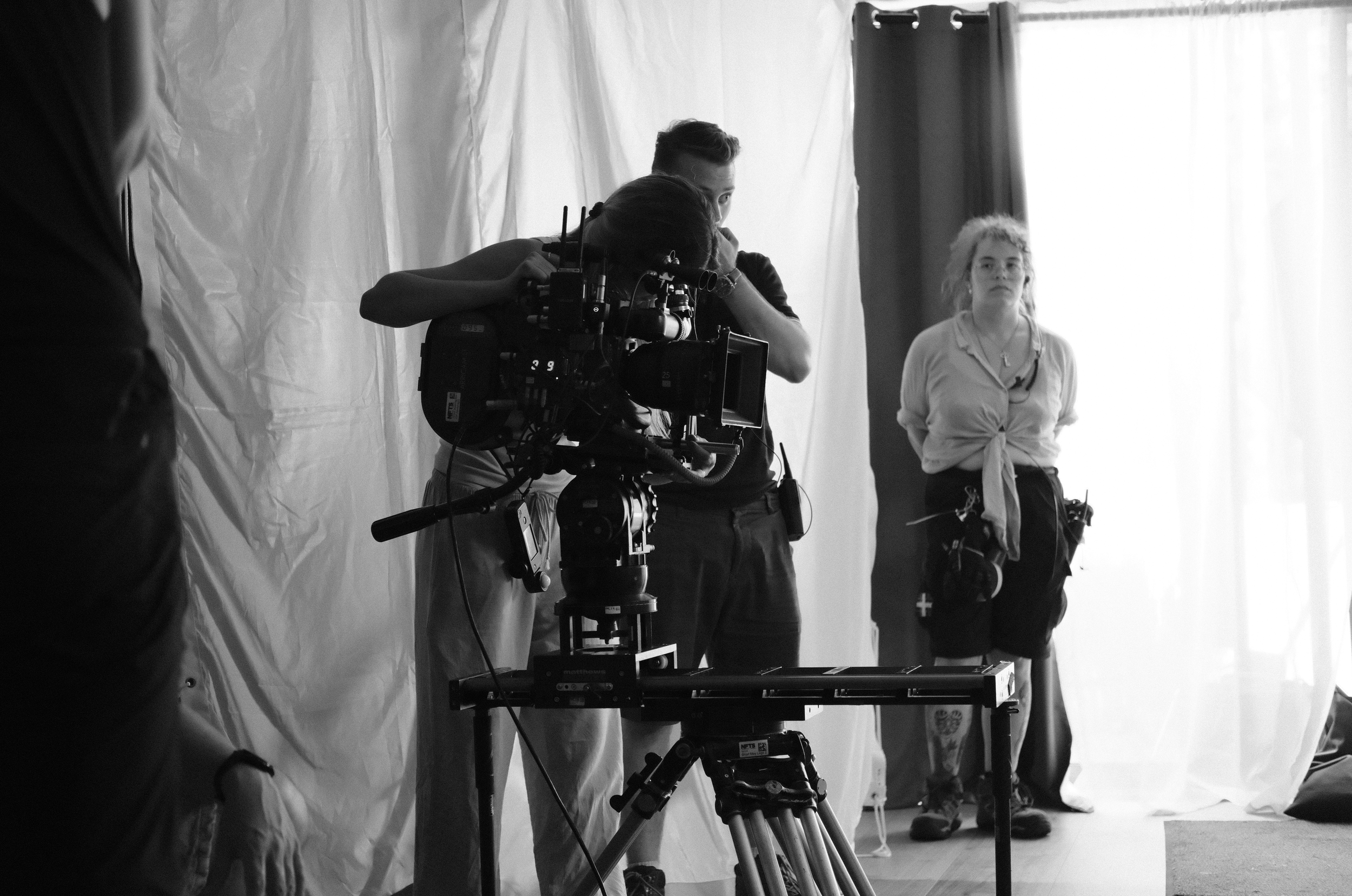 Black and white photo of a film set with a camera operator adjusting a camera, a woman standing in the background near a window with curtains.