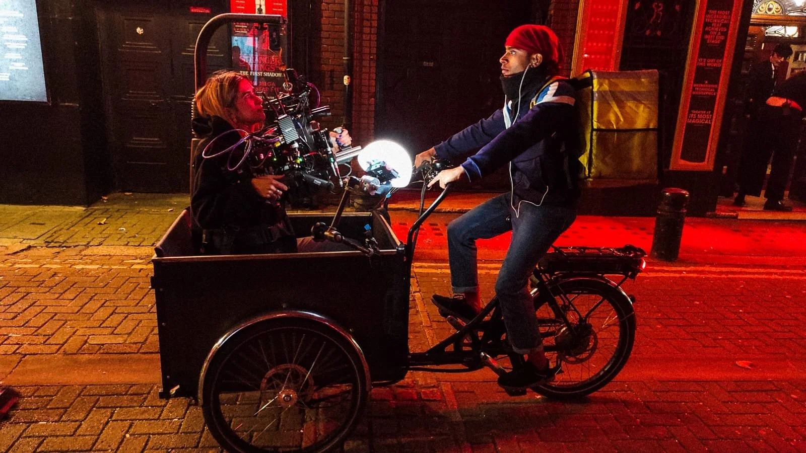 A man riding a bicycle with a cargo hold in the front is talking to a woman sitting inside the cargo area. The scene is lit with red and orange streetlights.