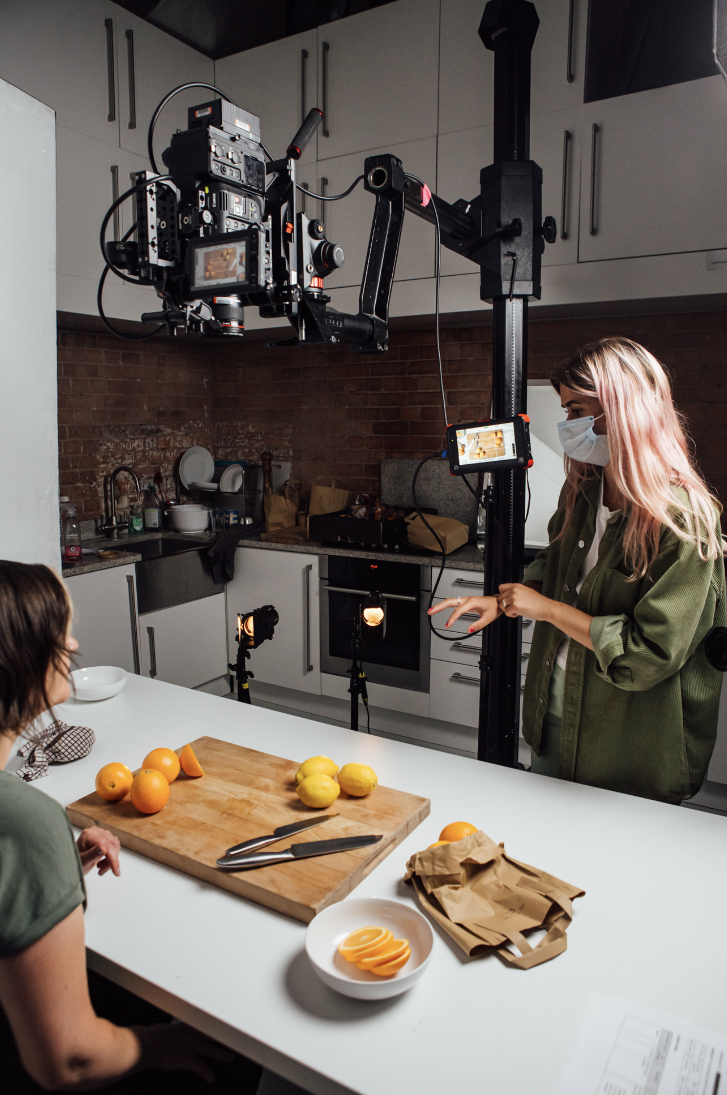 A woman wearing a face mask standing next to a camera rig in a kitchen, filming a woman sitting at a table with oranges and lemons, with studio lights directed at the table.