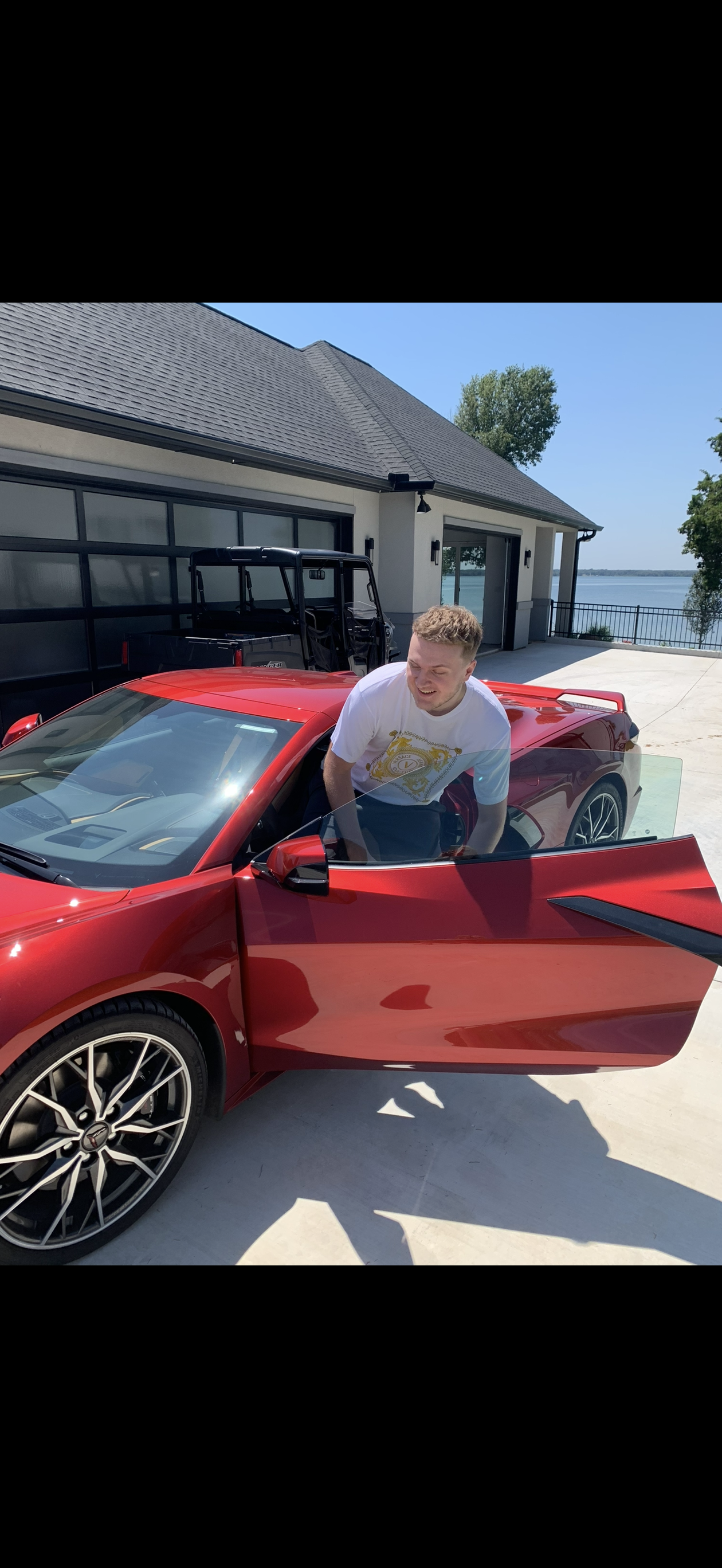 A man with light brown hair smiling inside a red sports car with the driver’s side door open, parked on a driveway near a modern house with large windows, overlooking a body of water under a clear blue sky.