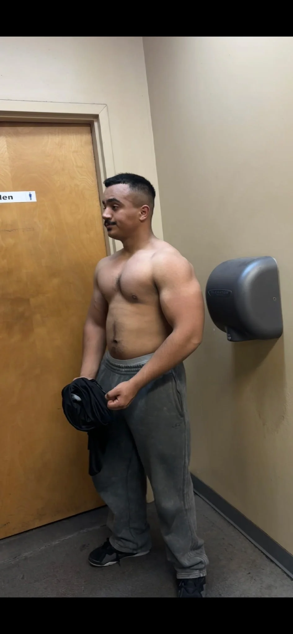 A shirtless man standing in a hallway, holding a black shirt in one hand, with gray sweatpants and sneakers. He is positioned in front of a closed wooden door with a men's restroom sign, and a paper towel dispenser on the wall beside him.