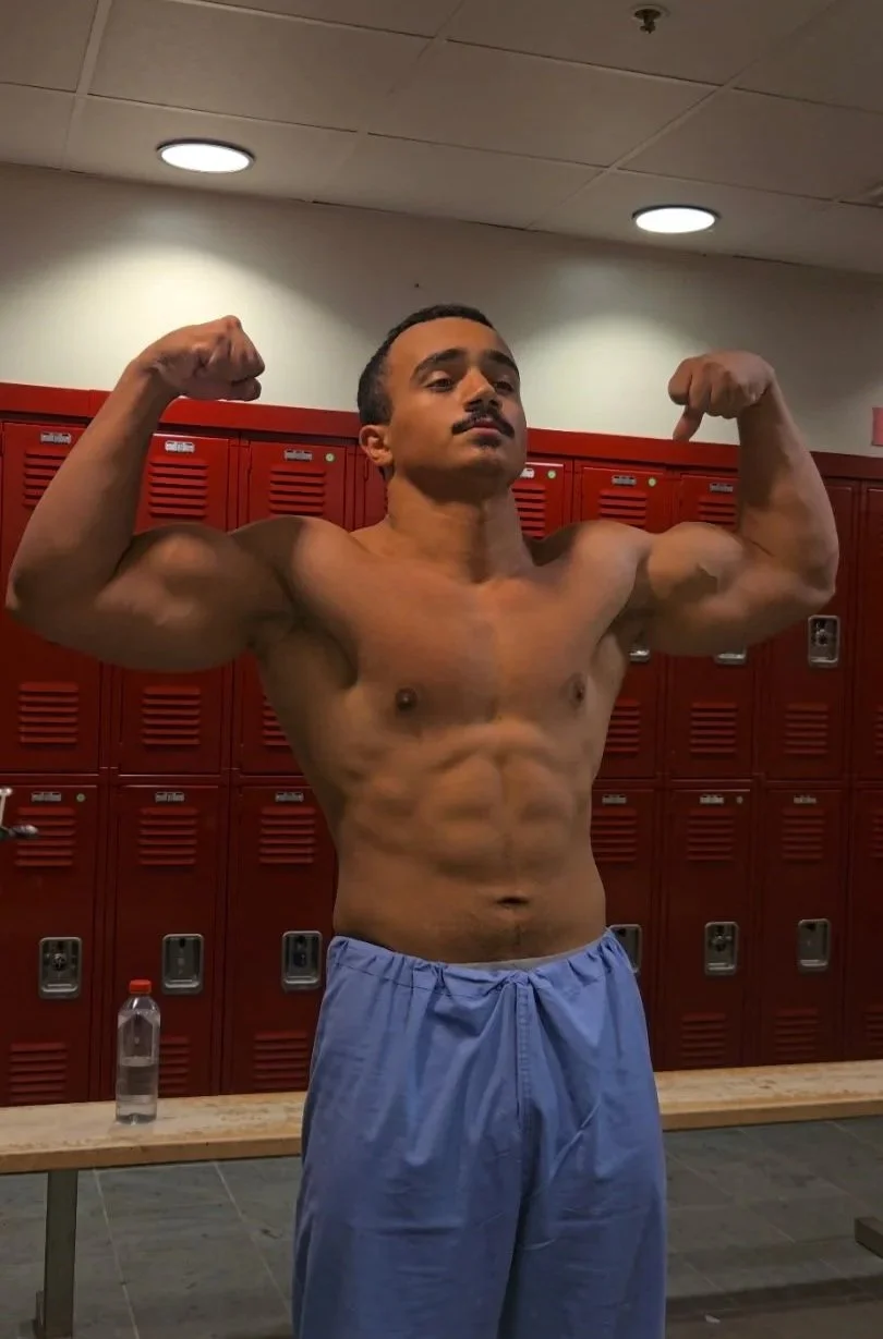 A muscular man with a mustache flexing his biceps in a locker room, wearing blue shorts, with red lockers behind him and a water bottle on a bench.