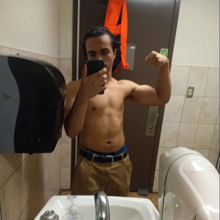 A shirtless man taking a mirror selfie in a bathroom, flexing his right arm. He is wearing khaki pants with a belt and has a serious expression.