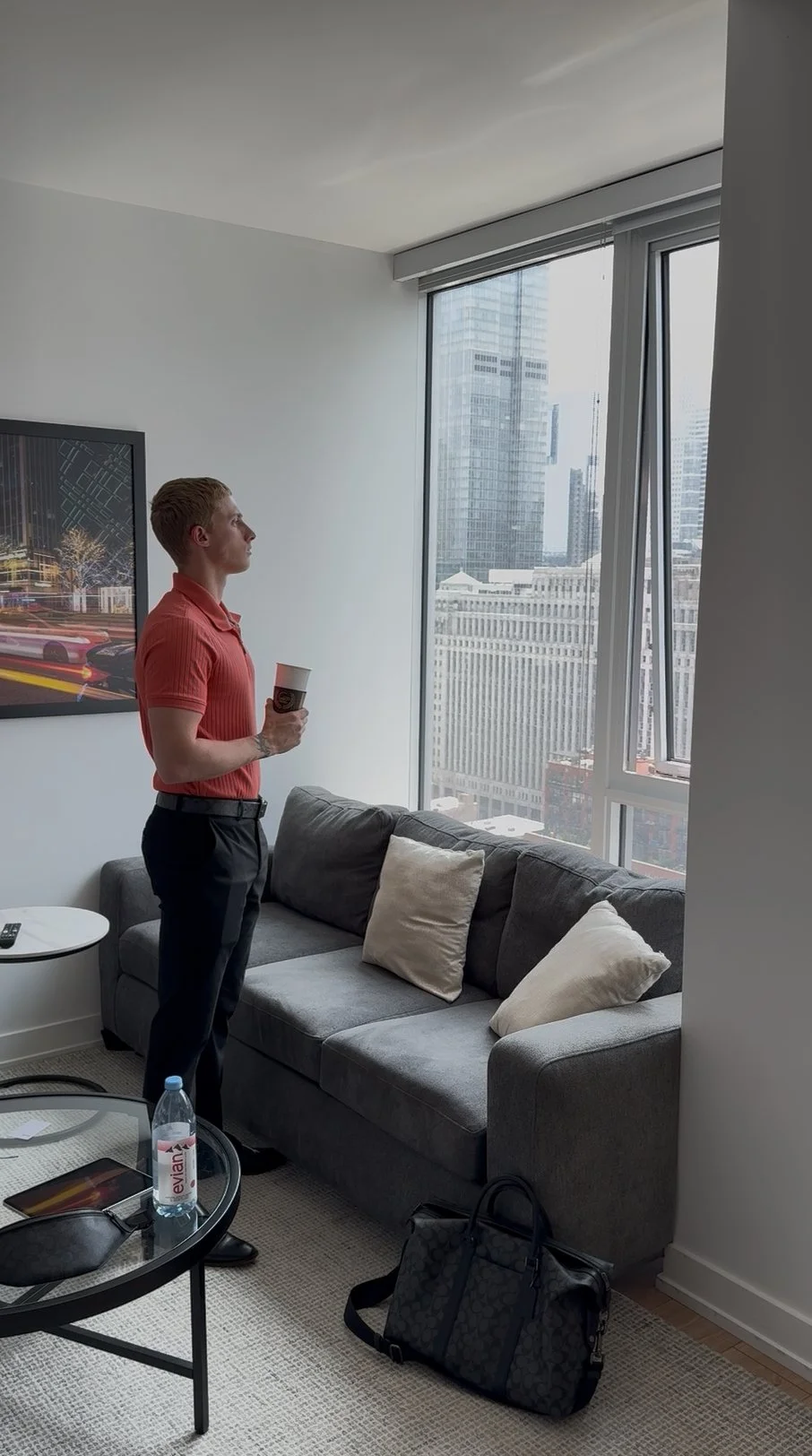 Man standing in living room holding coffee cup, looking out window at cityscape with tall buildings.