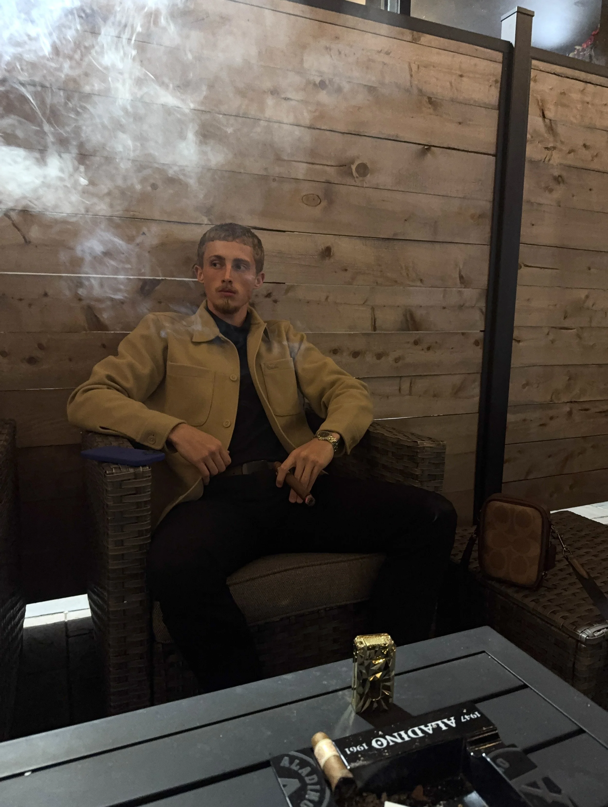 A man with short curly hair and a goatee sitting on a wicker chair against a wooden wall. He is wearing a beige jacket over a dark shirt. There is a glass of whiskey on the table in front of him, along with a cigar and a box labeled "1921 Ahlqvist". Smoke from the cigar is visible, rising near his face.