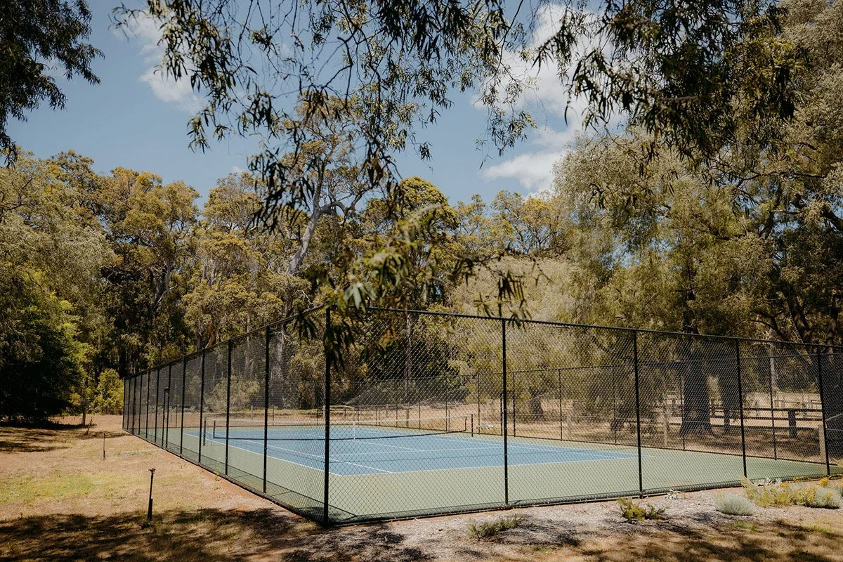 An outdoor tennis court surrounded by a black chain-link fence in a park with trees and a blue sky.