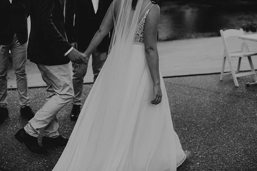 A bride in a flowing wedding dress holding hands with a groom or partner, standing outdoors near water with a bench in the background.