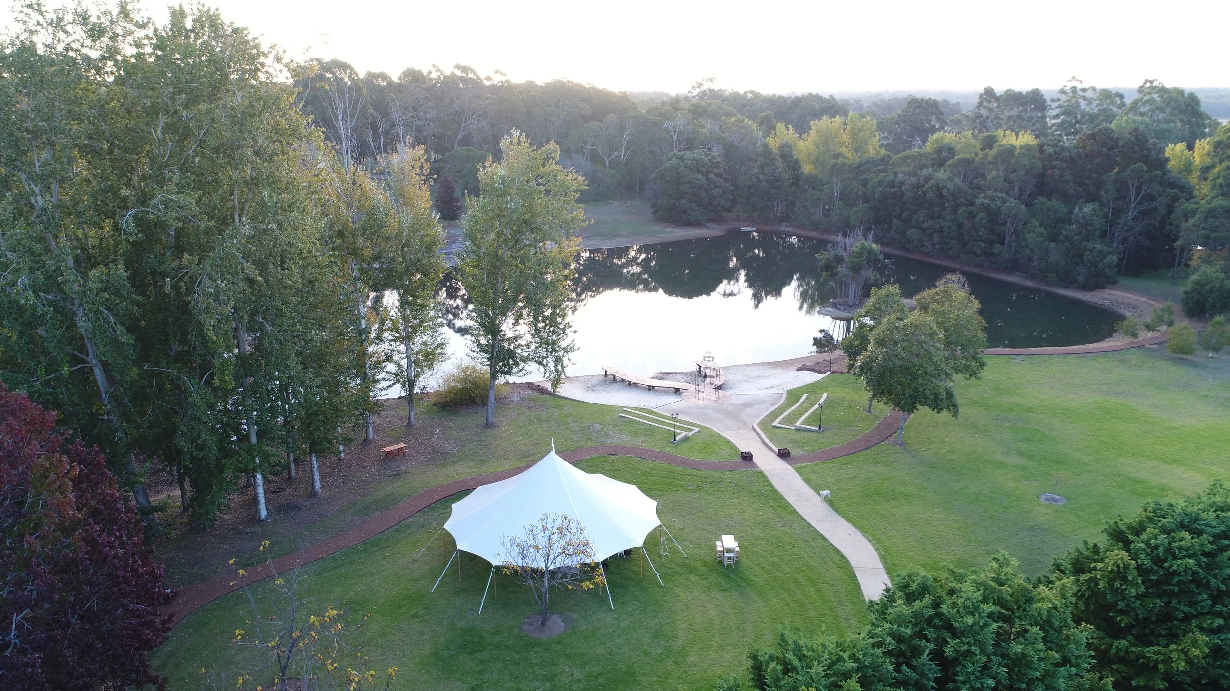 A park with a lake, surrounded by trees, with a white tent and pathways in the foreground.