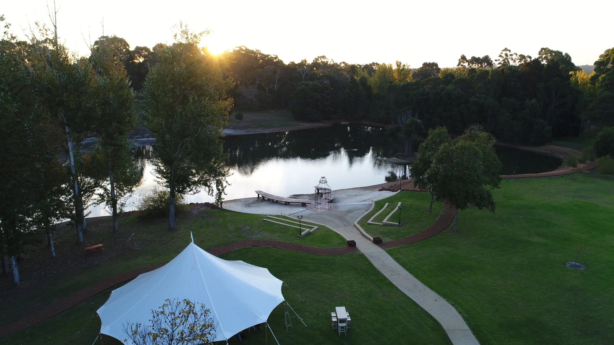 A park at sunset with a pond, trees, and a grassy area. There is a tent, a picnic table, and a small dock by the water.