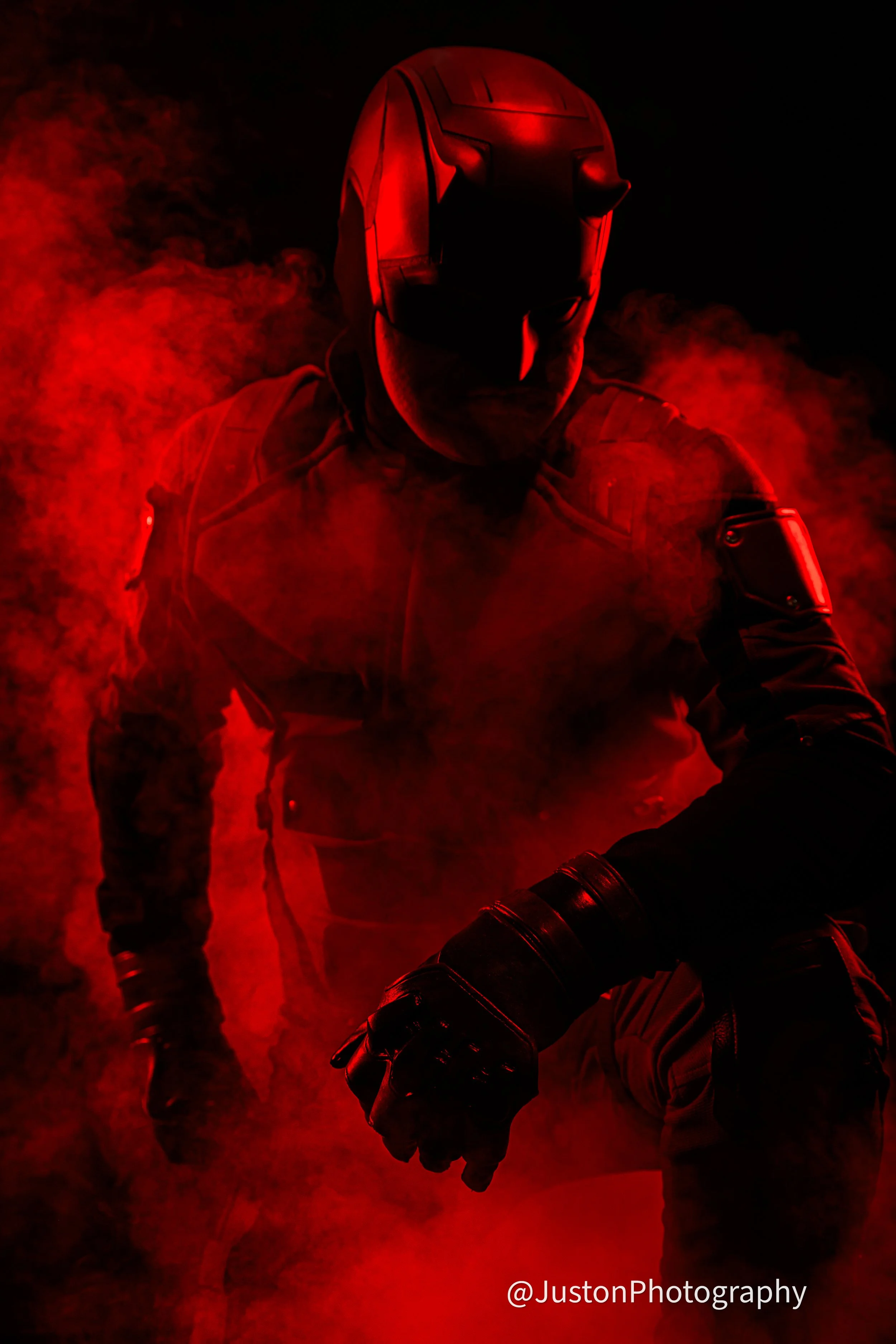 A person wearing a black tactical outfit and a mask, kneeling partially in red smoke with a dark background, illuminated by red lighting.