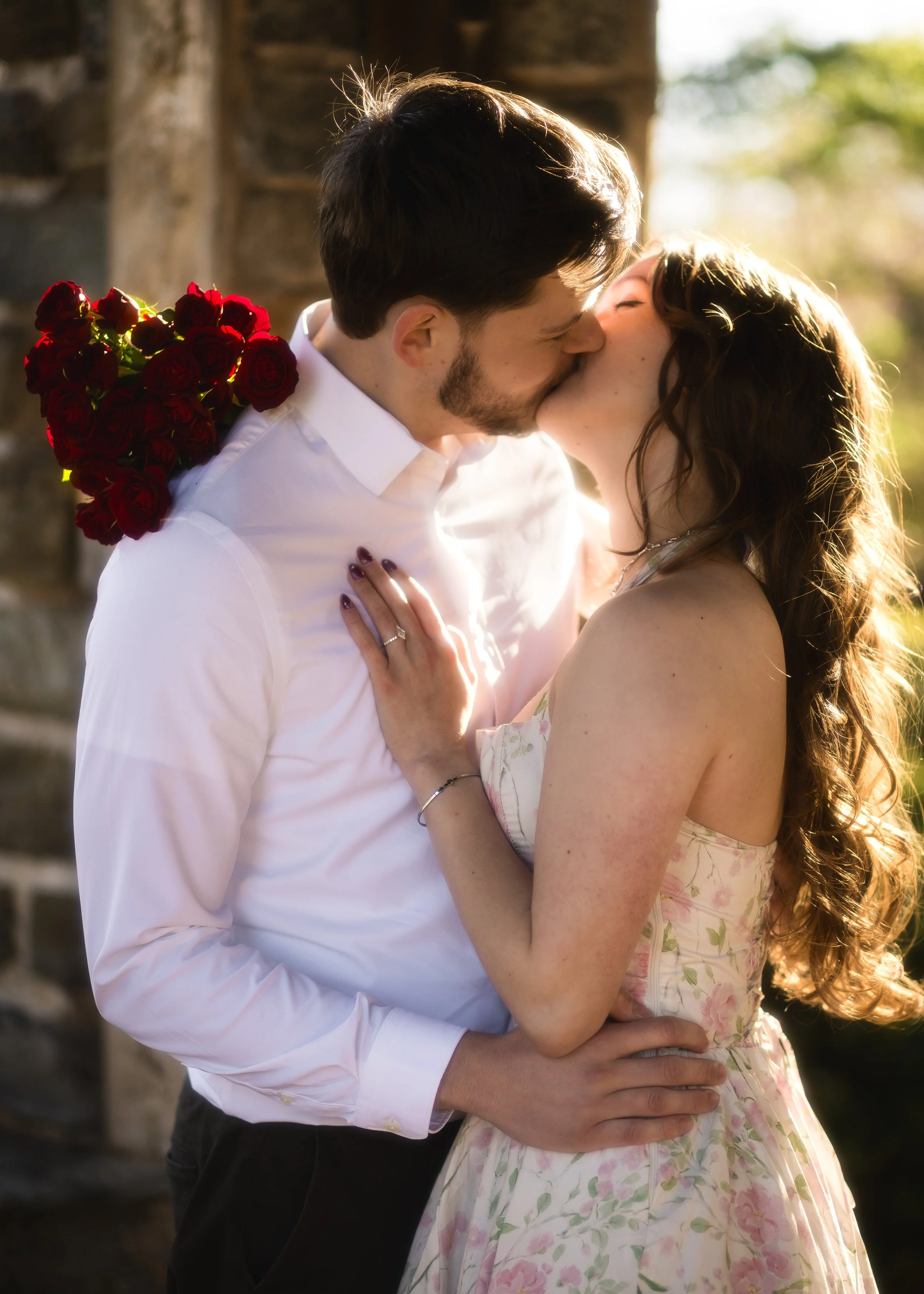 A couple kisses outdoors, with the woman wearing a floral dress and the man in a white shirt, holding a bouquet of red roses.