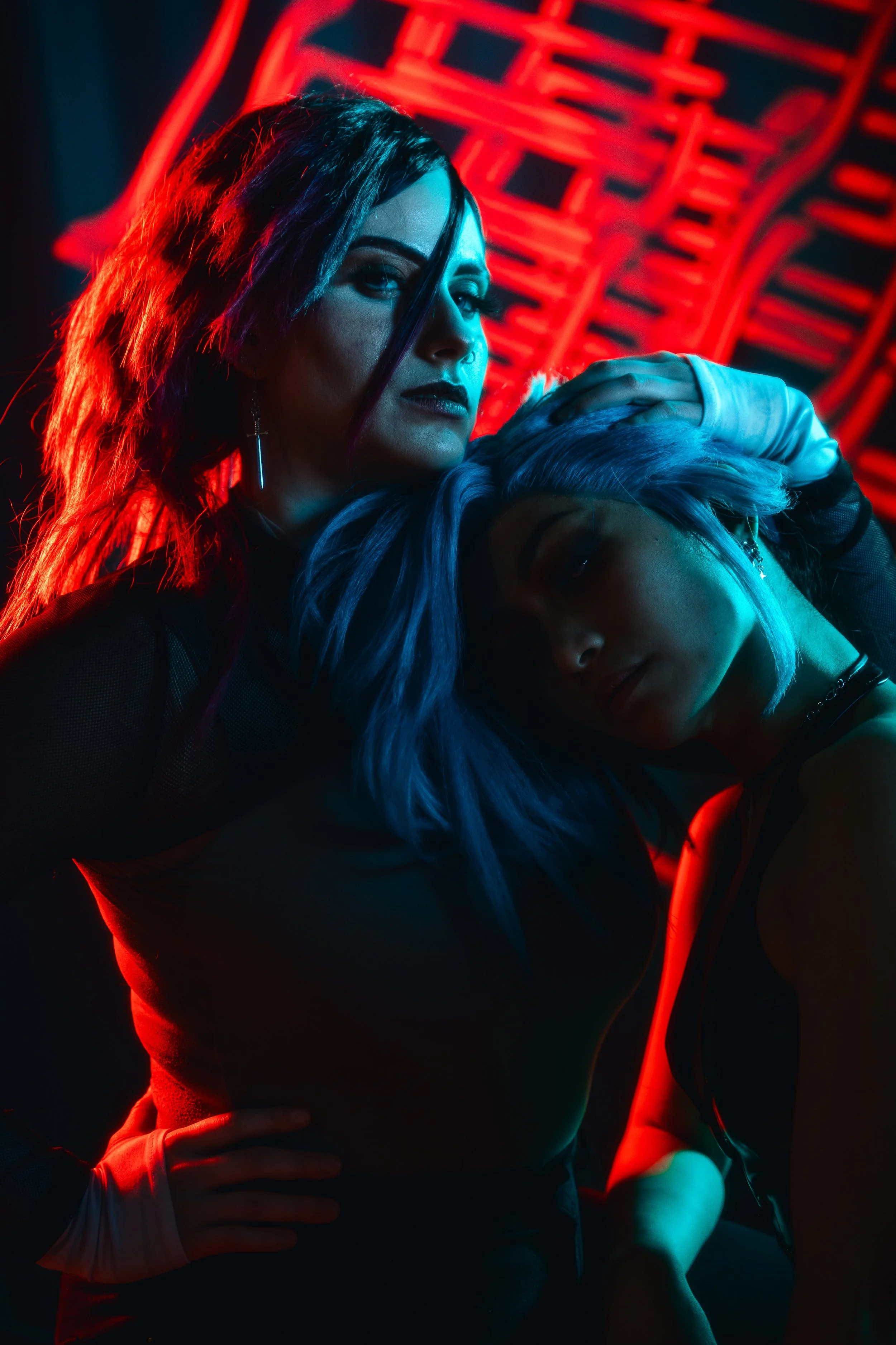 Two women with colorful hair, one with purple and blue hair and the other with blue hair, posing in a dark setting with red and blue lighting.