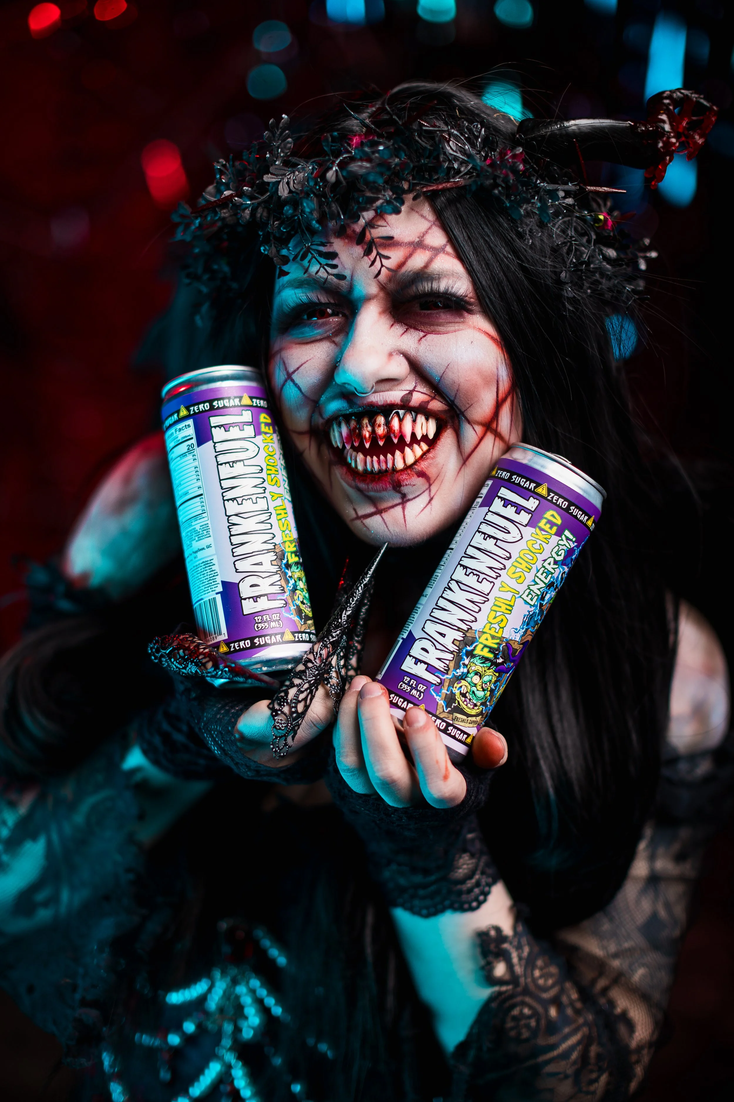 Person dressed in a vampire or gothic costume with face makeup, fangs, and a black lace headpiece, holding two cans of Transfusion energy drink, smiling under colorful lights.