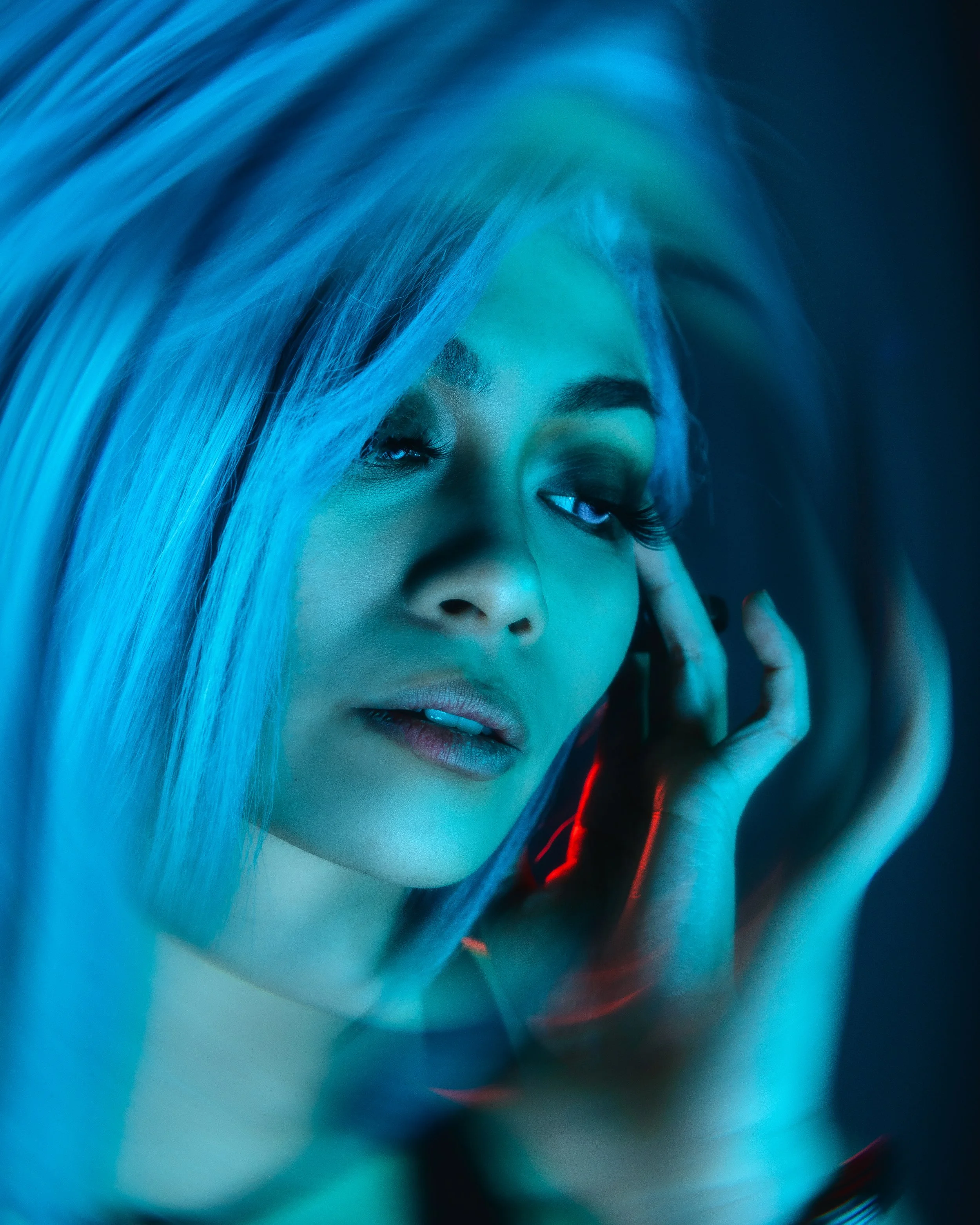Close-up of a woman with blonde hair, wearing makeup, with her hand near her ear, illuminated by blue and red lighting.
