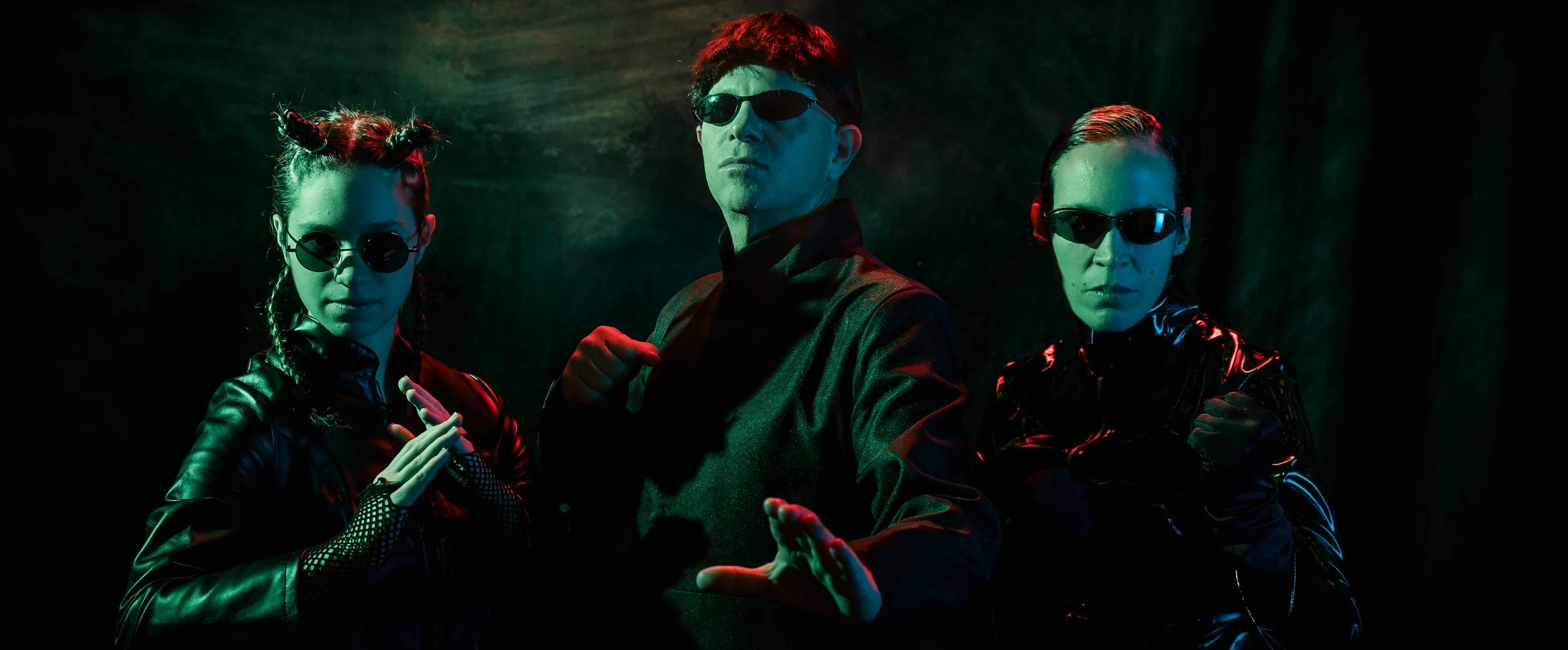 Three individuals dressed in black, wearing sunglasses, posing against a dark background with neon lighting effects.